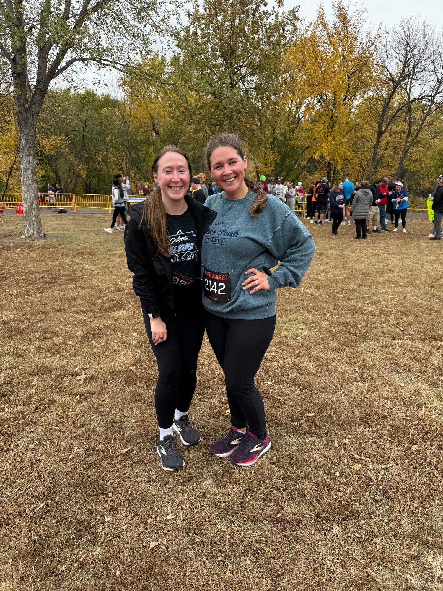 Two women smiling together outdoors at fall 5K race event wearing race bibs.