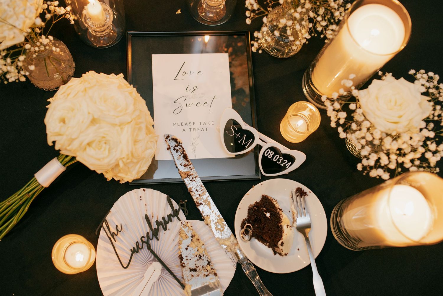 Wedding dessert table with cake slices, white roses, candles, and Love is Sweet sign display.