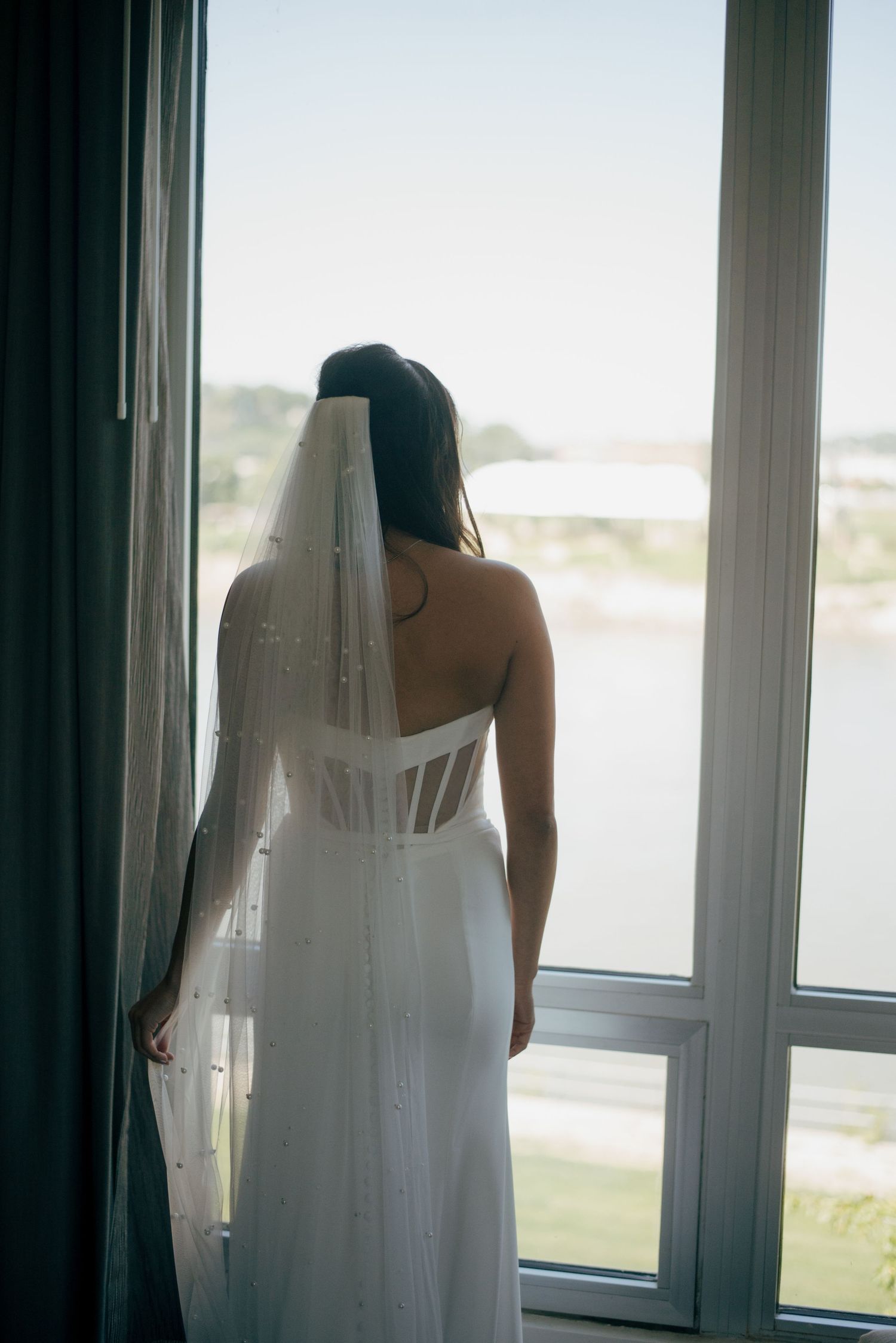 Bride in white corset wedding gown and veil stands gazing out large window overlooking water.