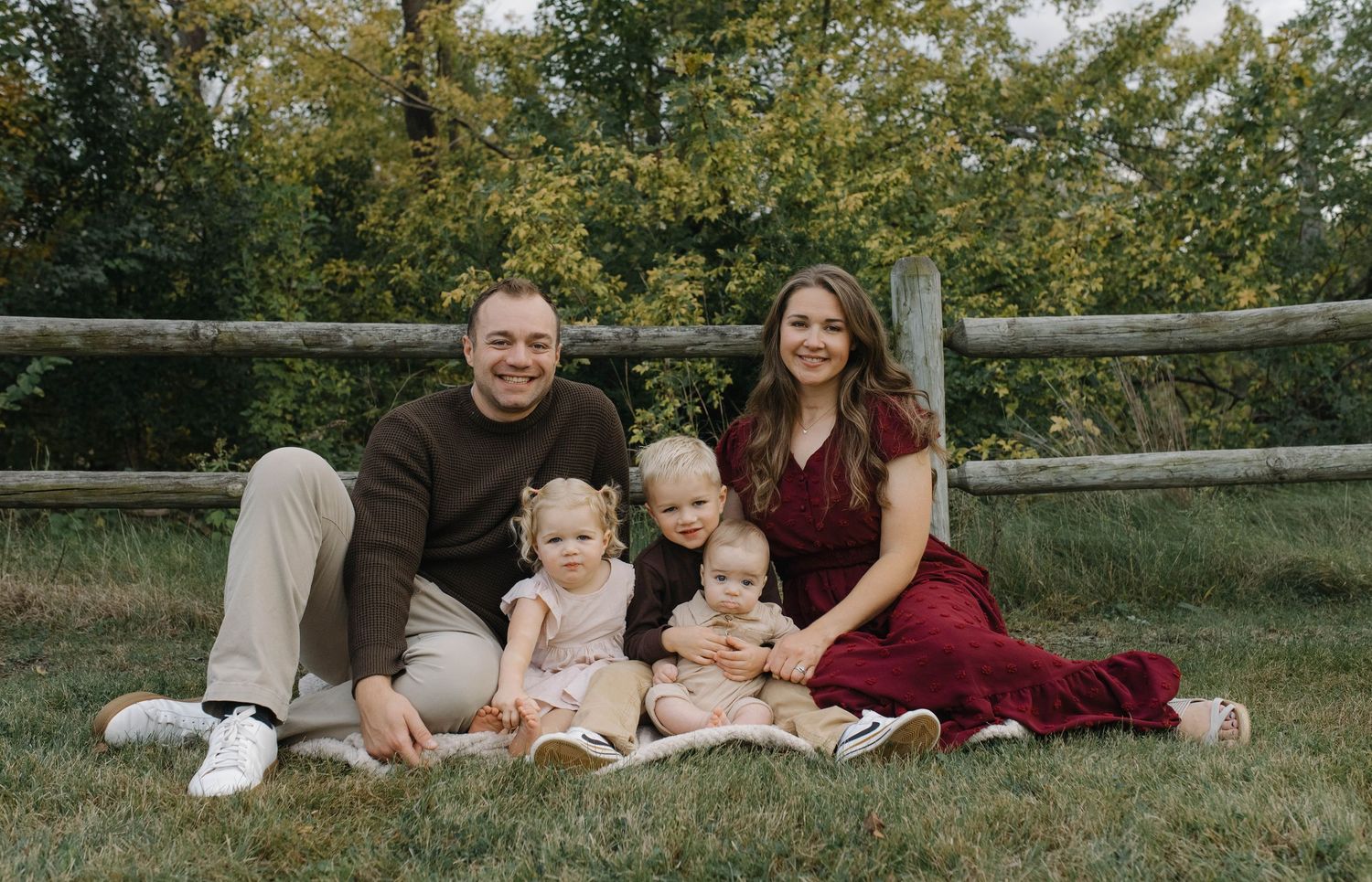 Happy family of five with three young children sitting on grass in front of a wooden fence for a portrait.