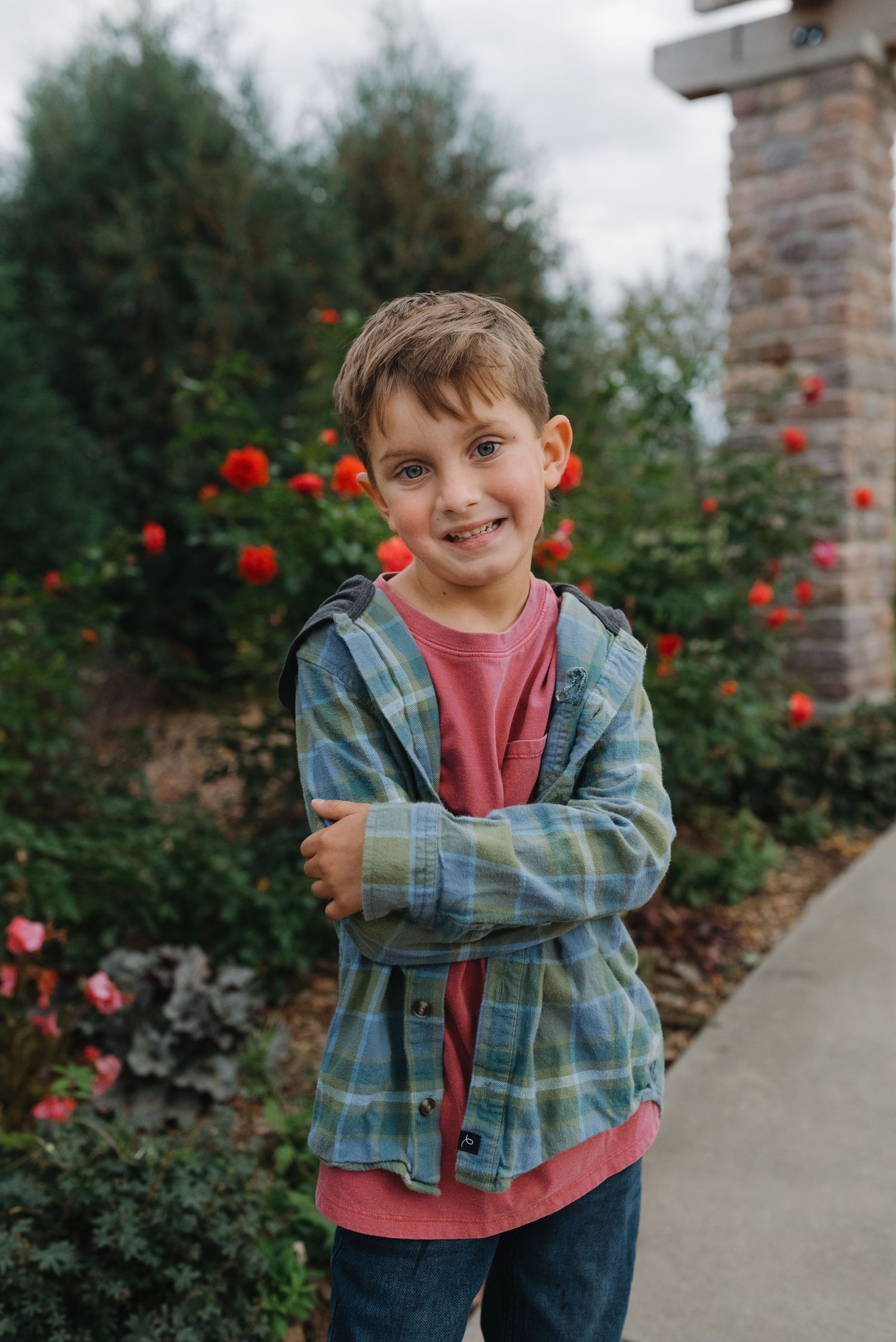 Young boy in a plaid jacket smiling with arms crossed in a garden with vibrant red roses.