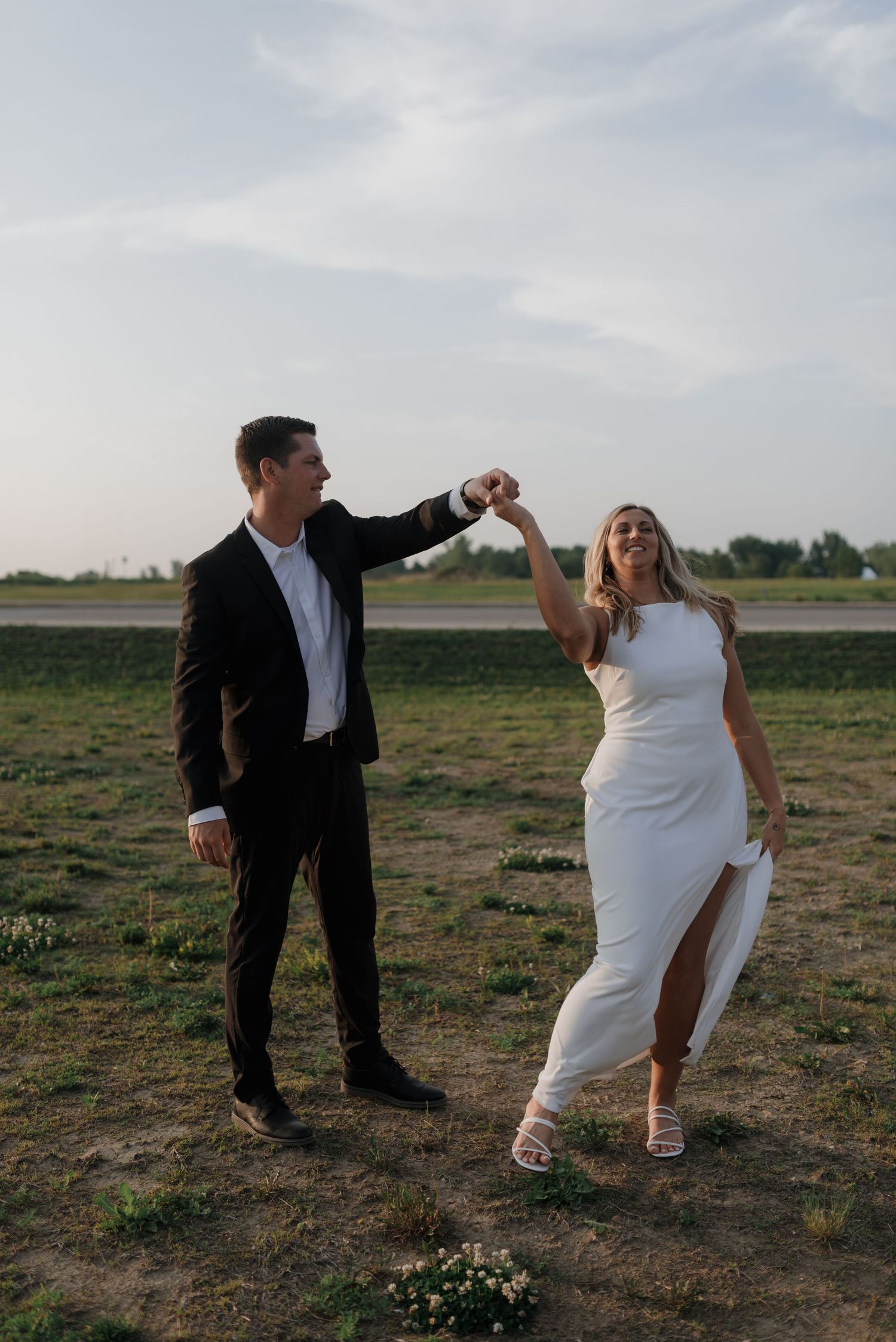Couple dancing and laughing outdoors at sunset, man twirling smiling woman in white slit dress.
