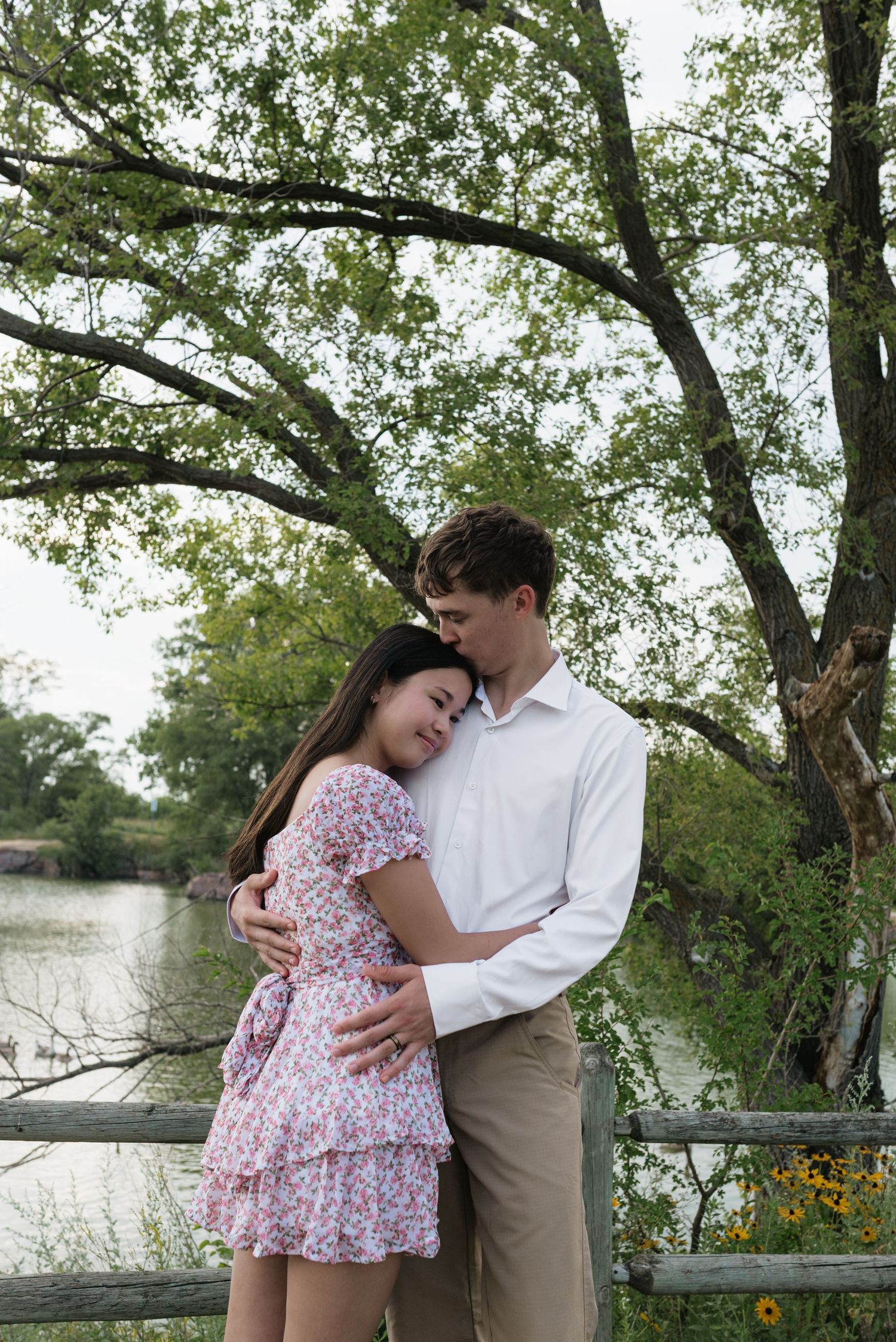 A couple shares a tender embrace near a wooden fence by a peaceful lake surrounded by greenery.