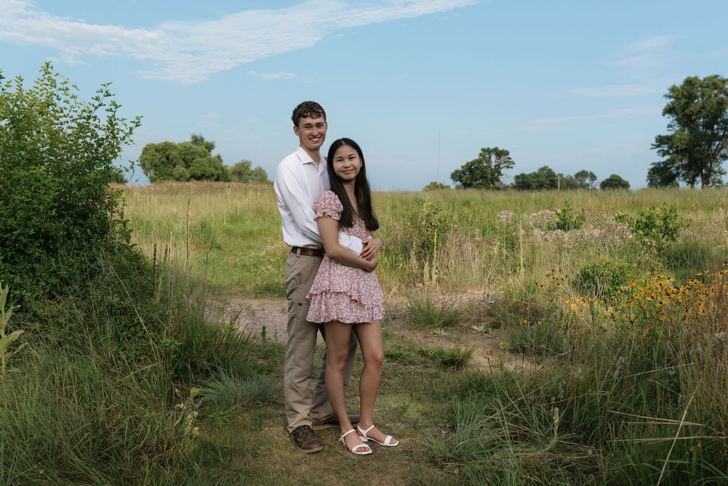 Couple embracing on a dirt path in an open field with tall grass and blue sky.
