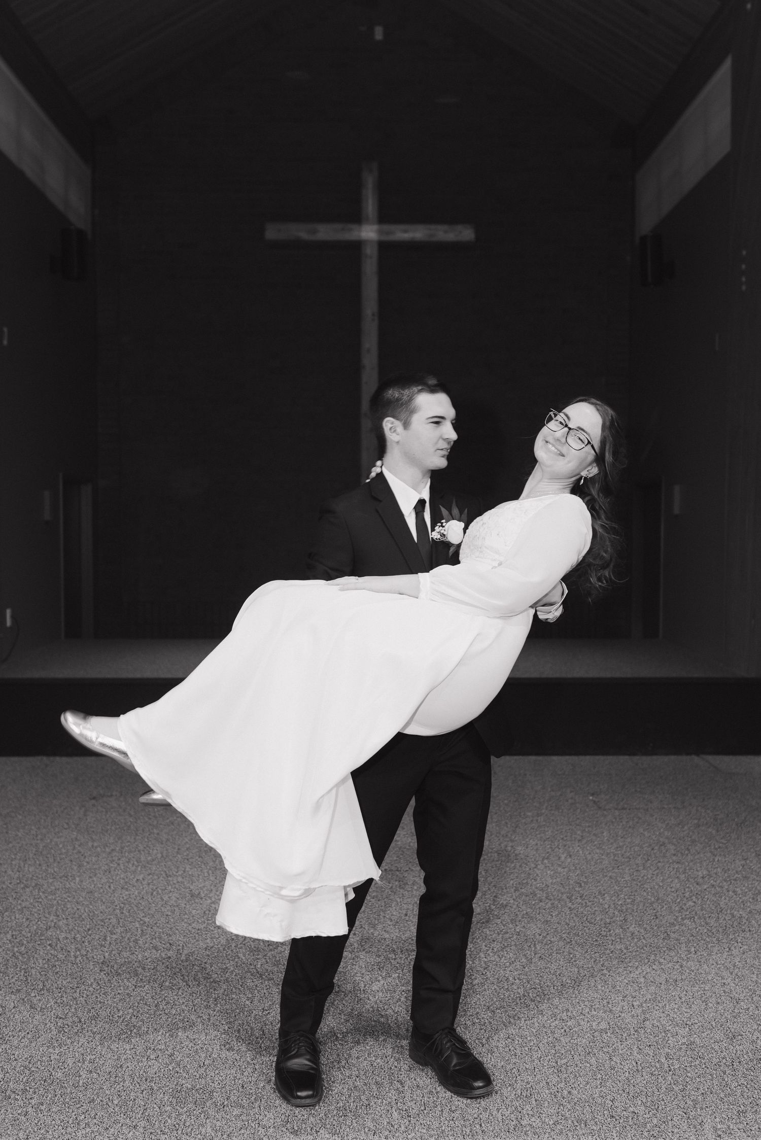 Groom in black suit carries smiling bride in white dress inside church with cross in background.