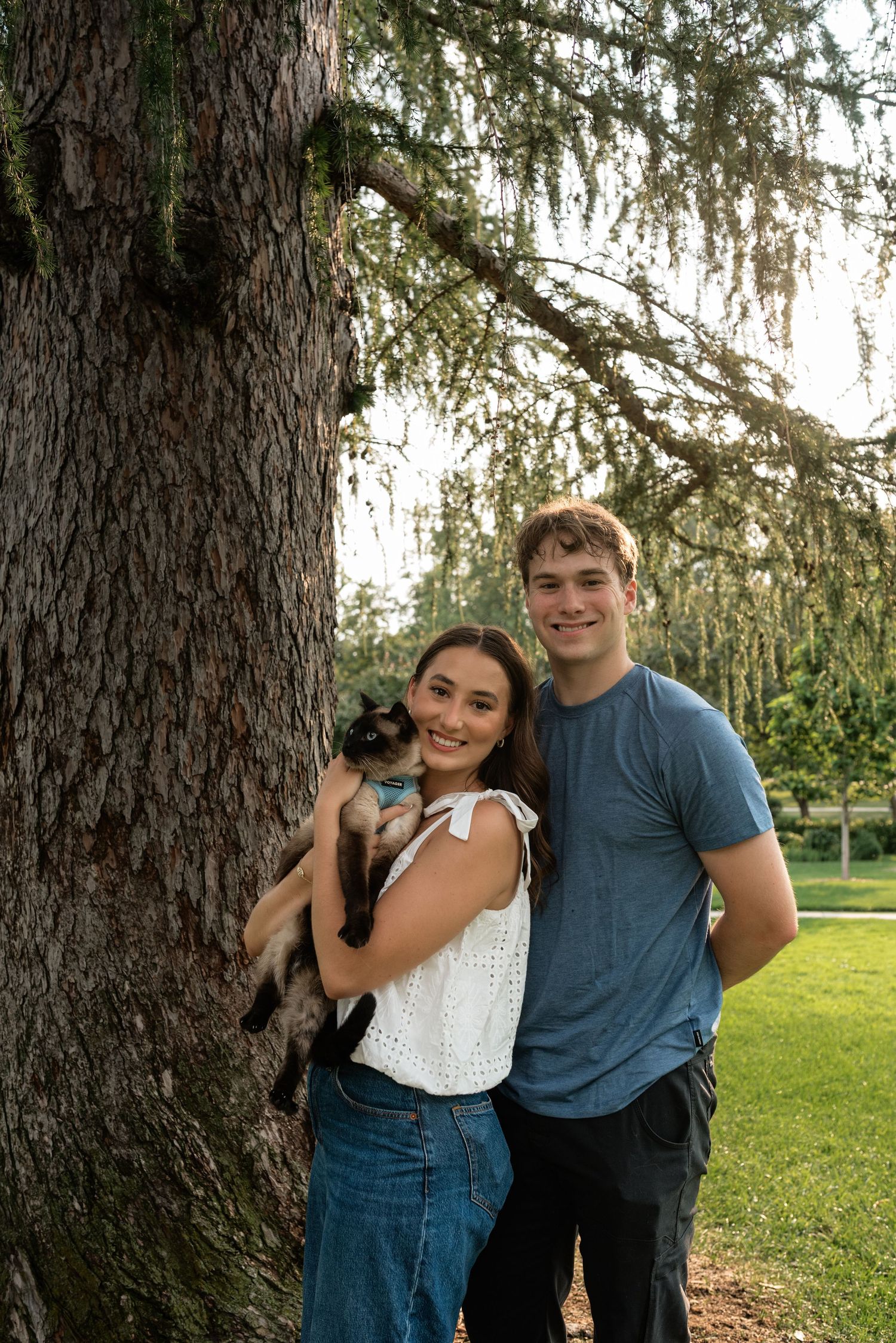 Young couple smiling outdoors by a large tree, woman holding a Siamese cat, man in blue t-shirt beside her.