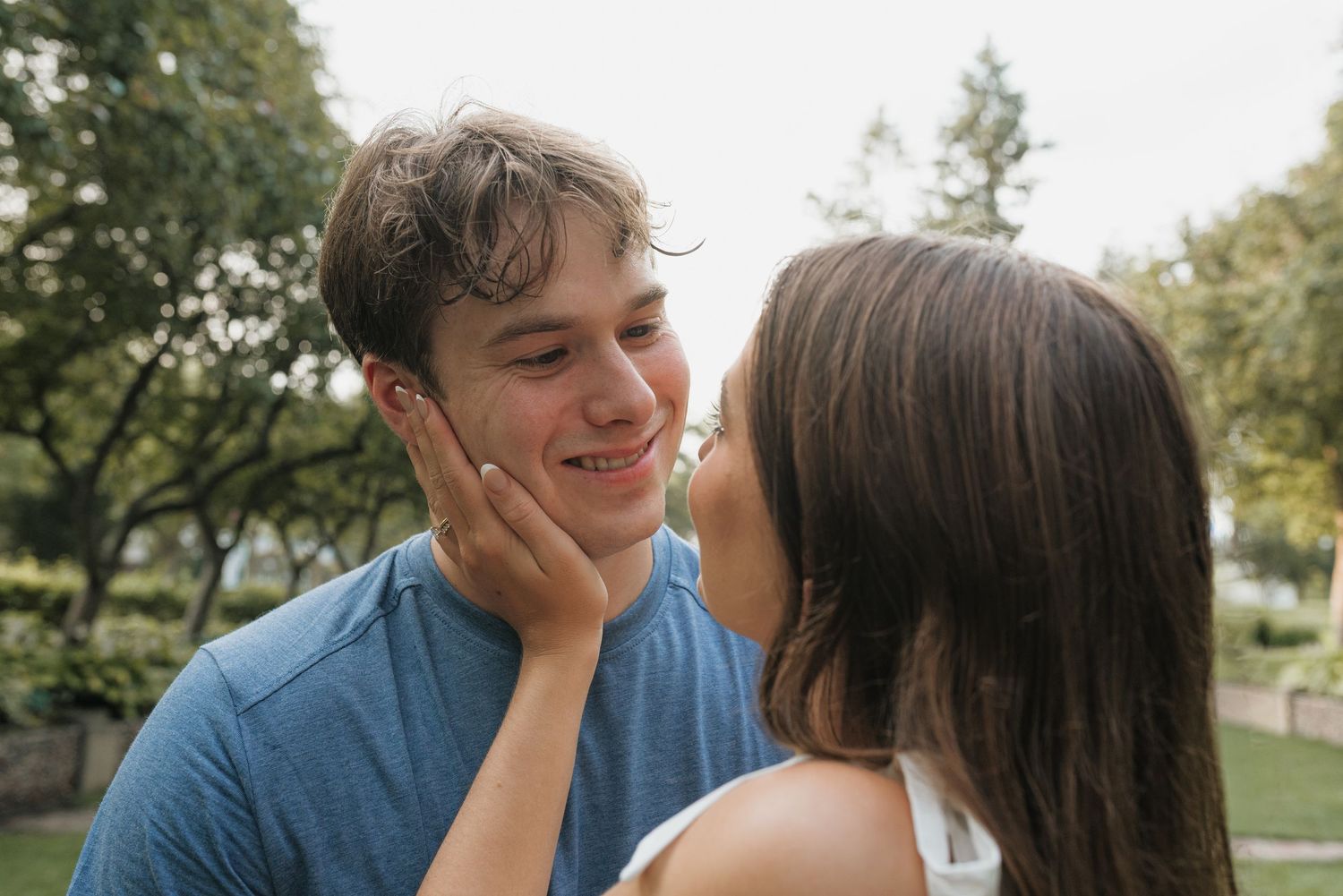 A young couple shares a romantic moment outdoors, with a woman gently touching a smiling man's face in a park.