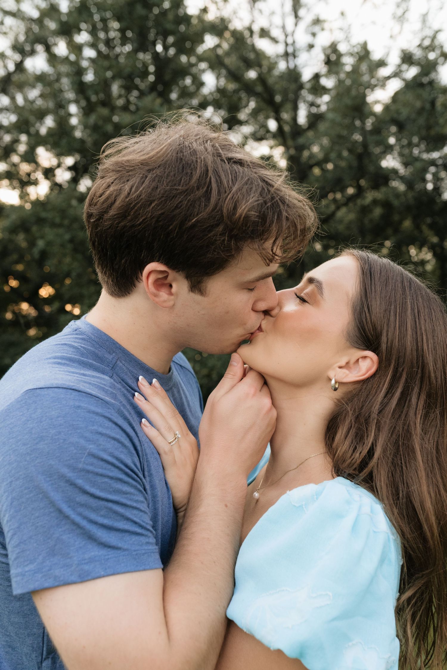 A young couple shares a romantic kiss outdoors, with lush green trees in the background at sunset.