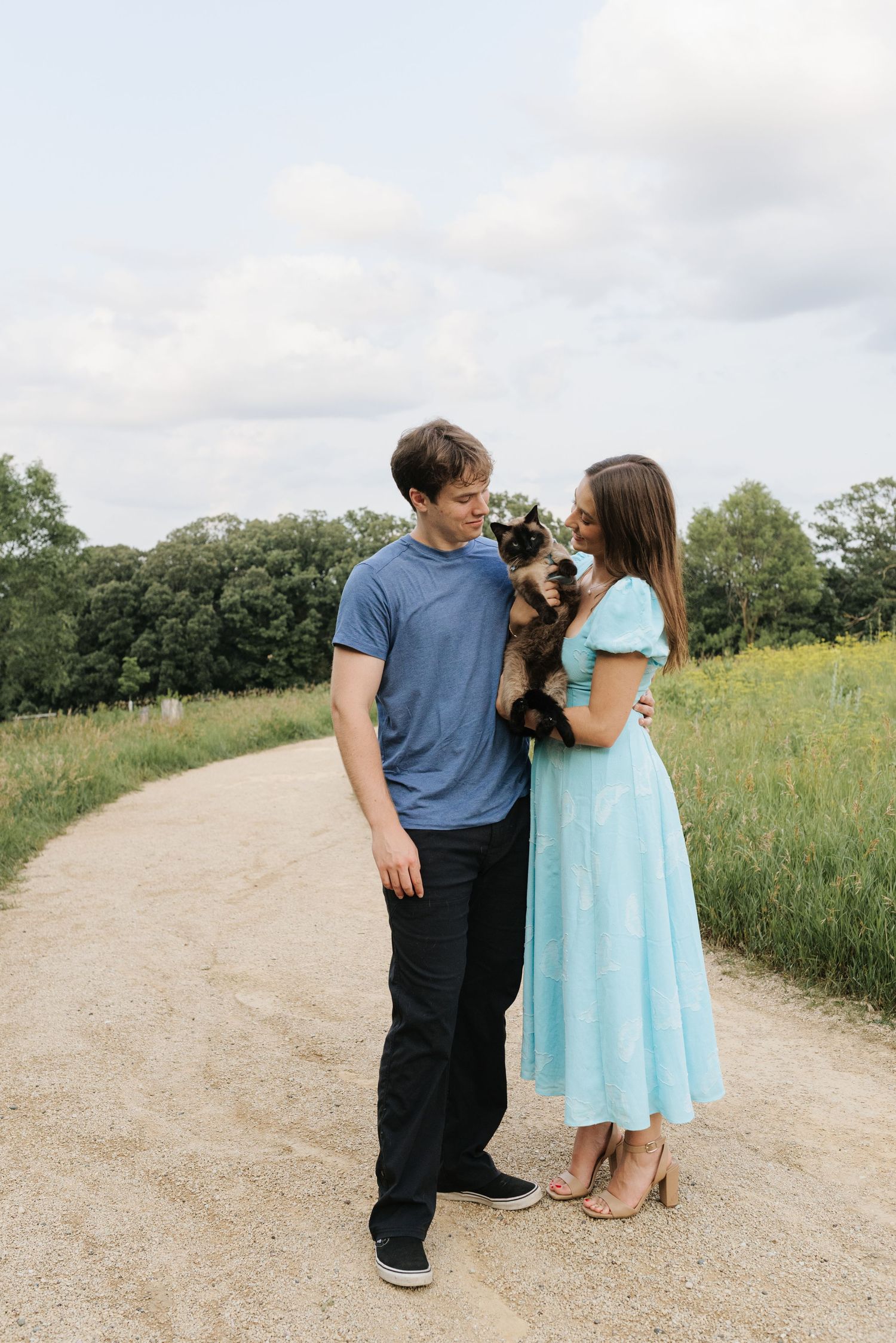 A couple stands on a dirt path holding a cat, surrounded by green fields and trees under a cloudy sky.