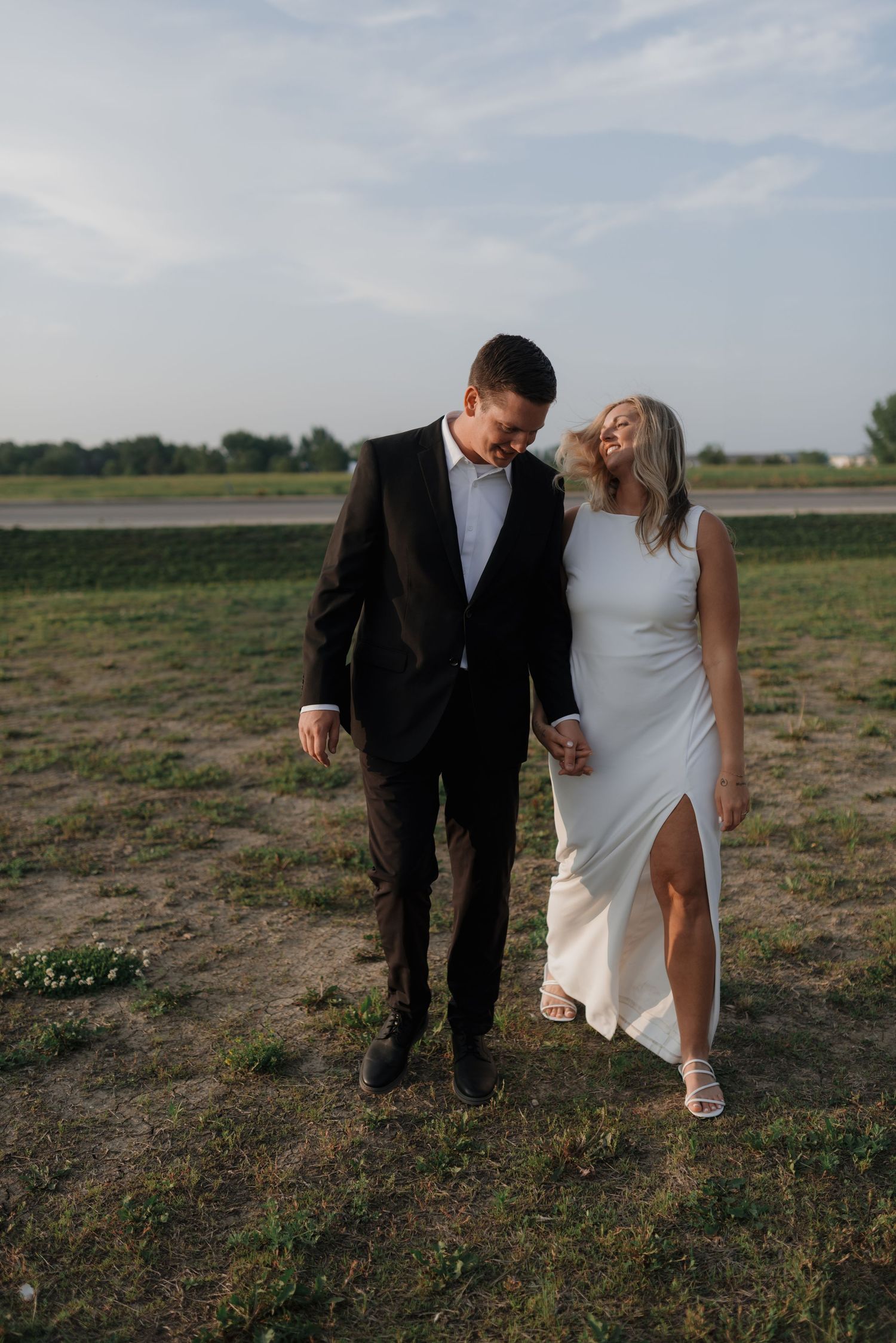 Bride leaning on groom while walking hand in hand through a scenic outdoor field.