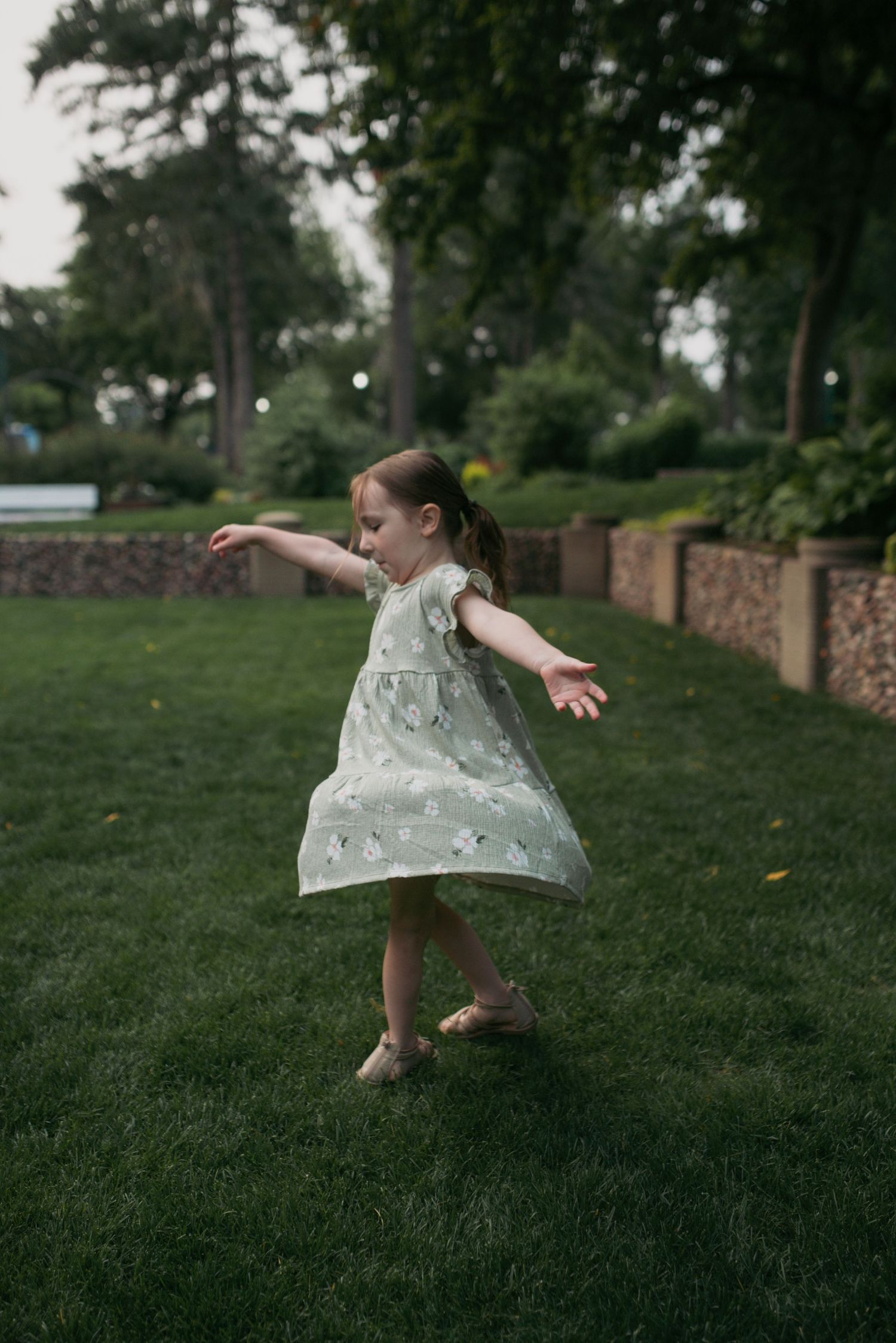 Young girl in floral green dress twirling joyfully on lush grass in a park setting.