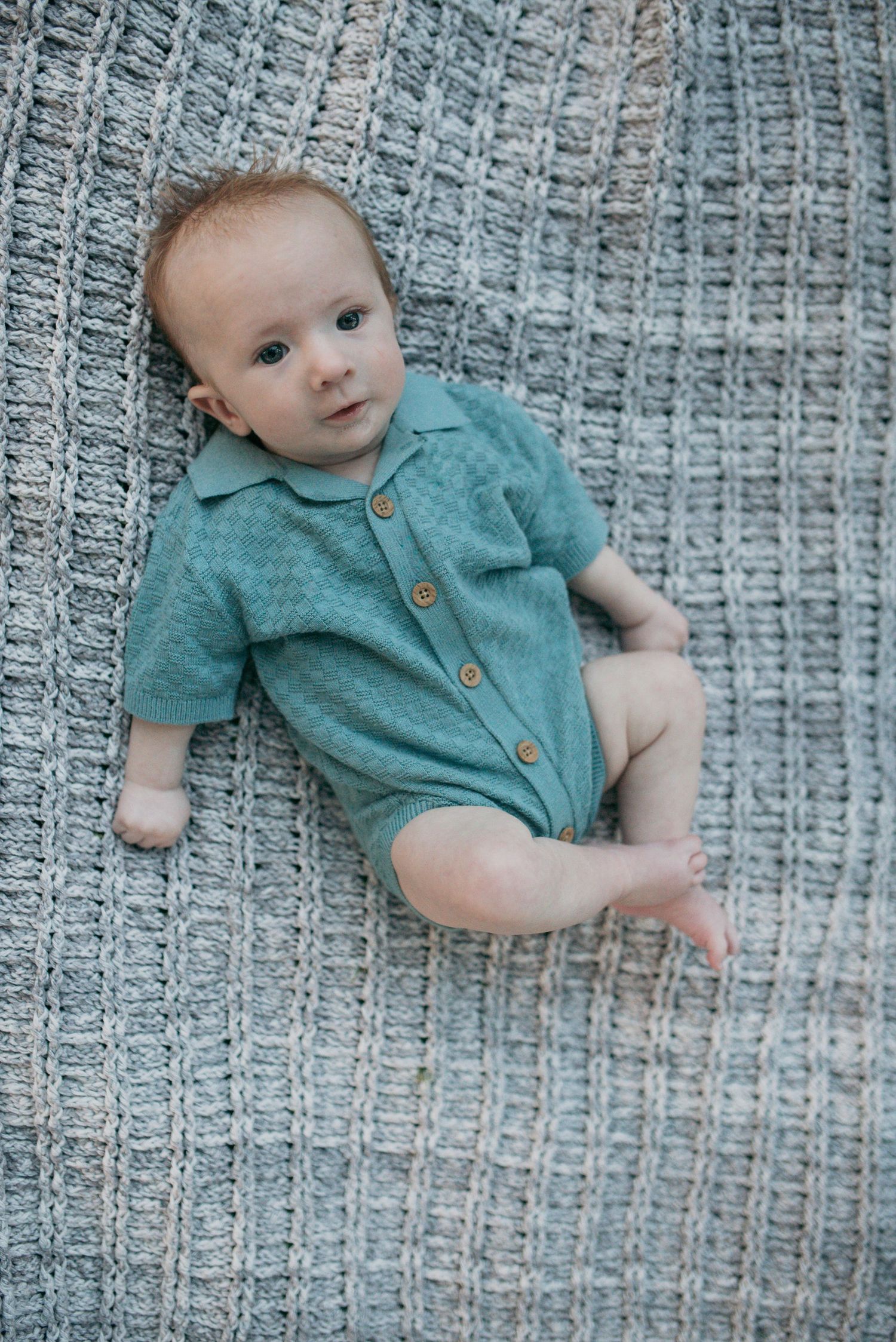 Cute infant wearing teal collared romper resting on textured gray knit blanket.