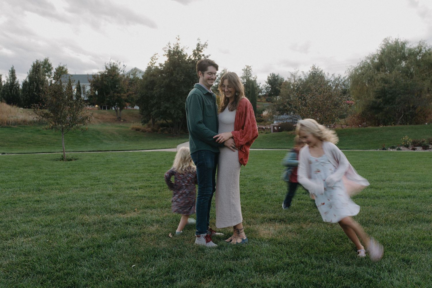 Couple standing on green grass while children play around them in a park.