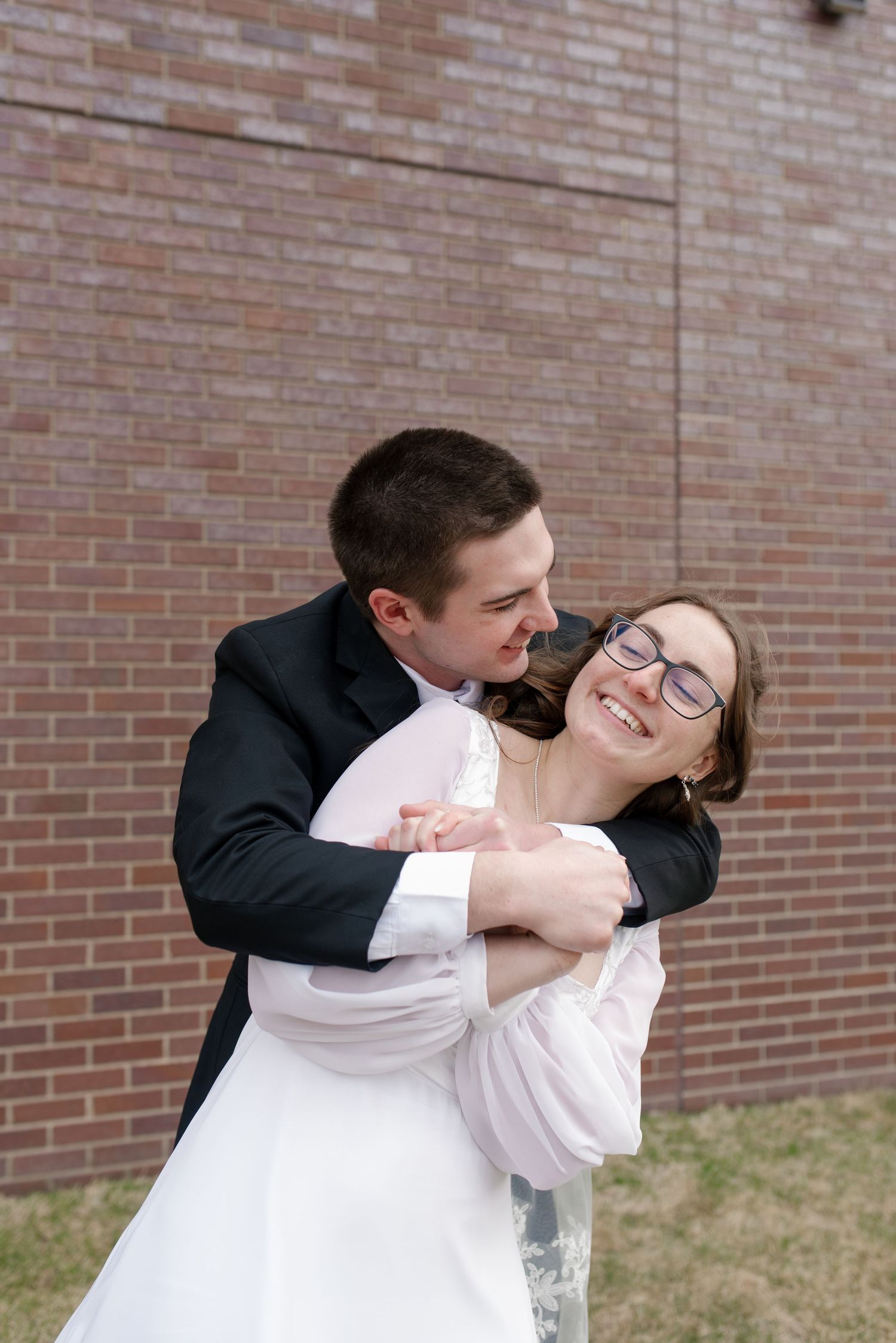 Groom joyfully embraces laughing bride in white dress and glasses against brick wall backdrop.