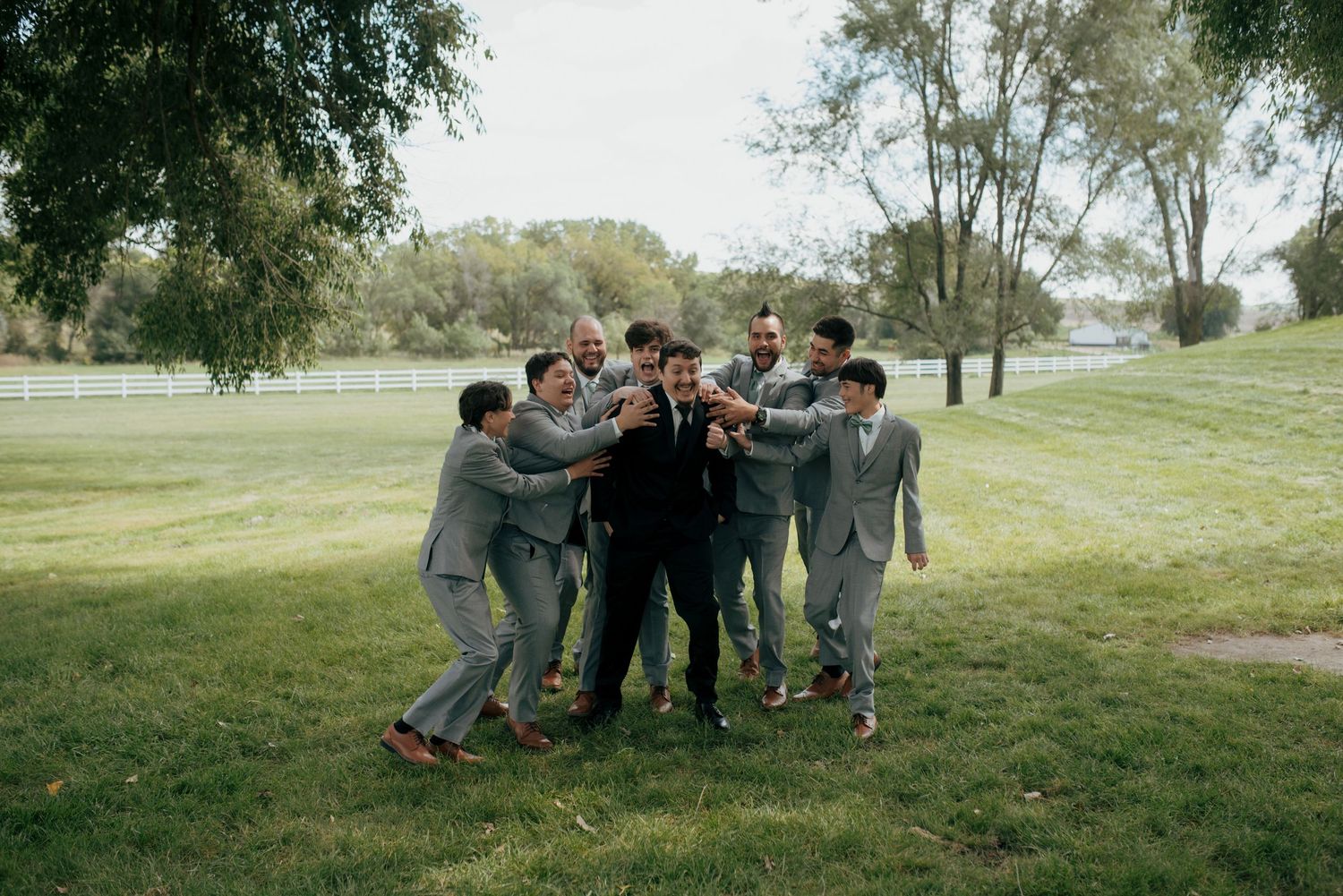 Groomsmen in grey suits huddle together celebrating on green lawn with trees in background.