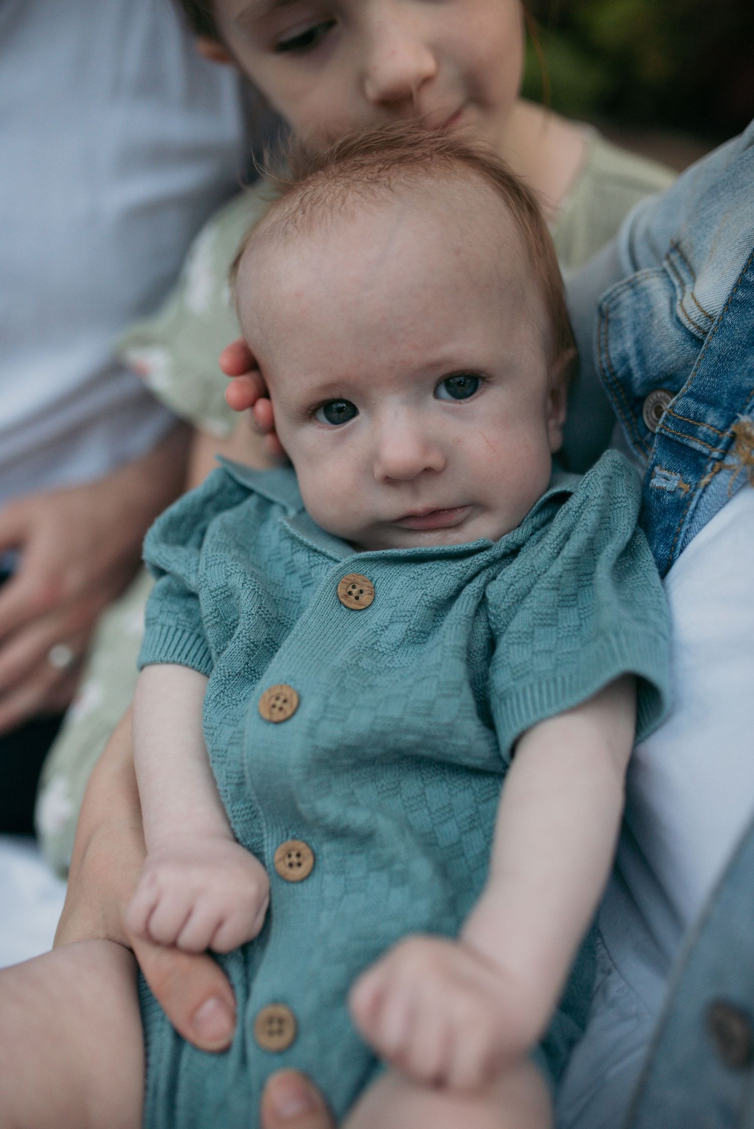 Close-up of an adorable baby in a teal knit button romper being held by family.