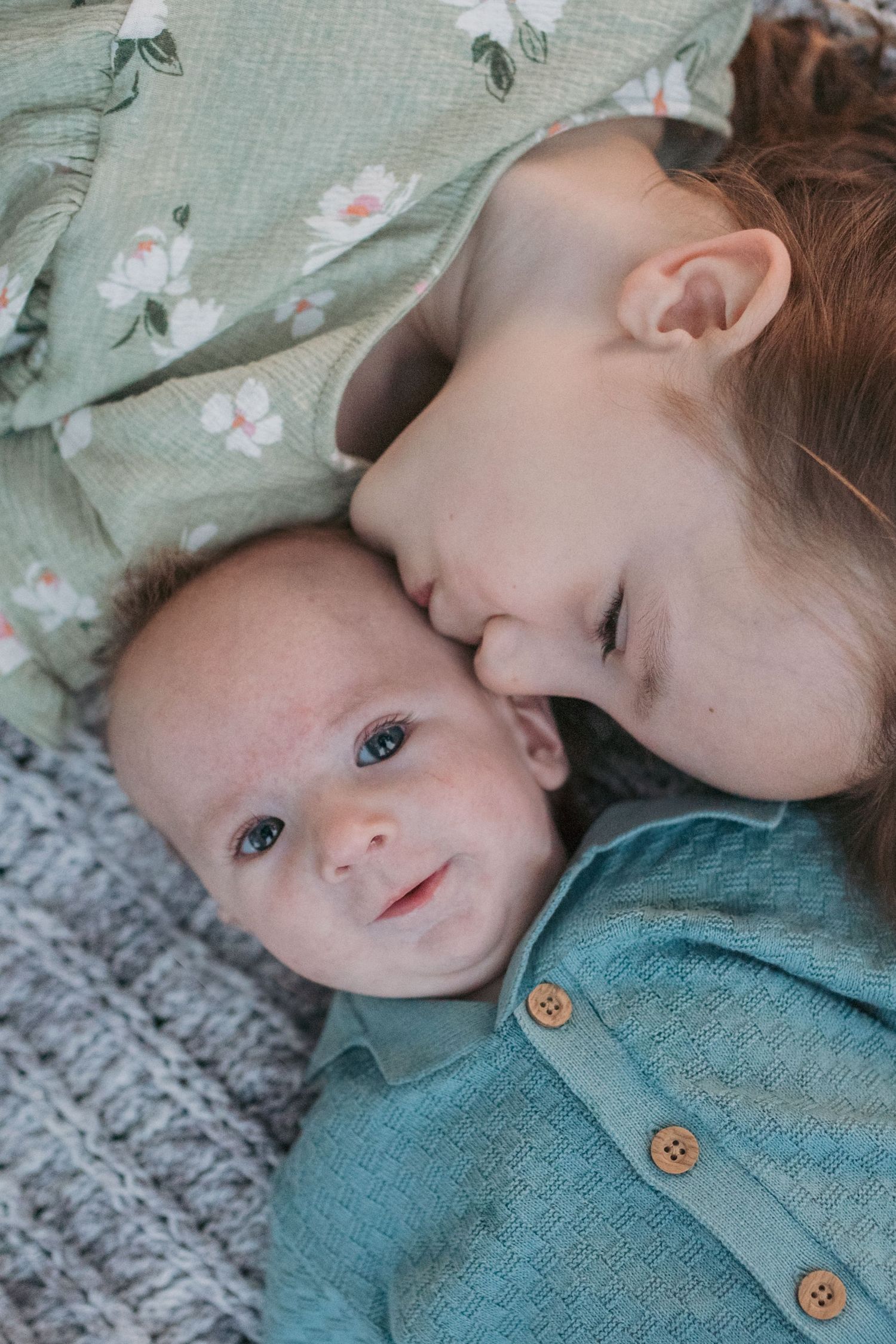 Loving older sister gently kissing newborn baby sibling on cheek on gray blanket.