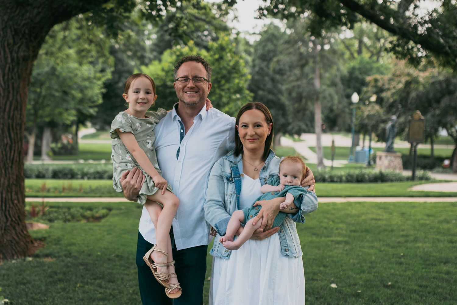 Smiling family of four standing in a lush green park, parents each holding a young child.