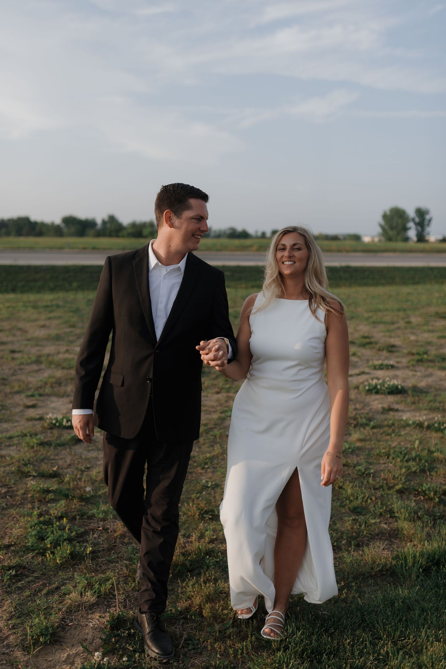 Bride and groom walking hand in hand through a green field at golden hour in elegant wedding attire.