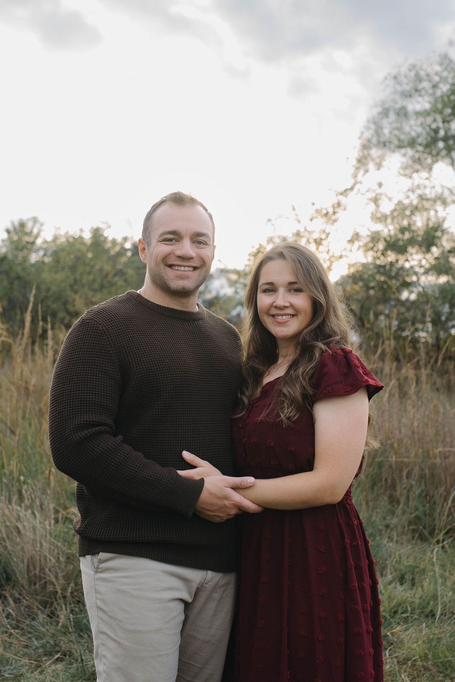 A smiling couple holds hands in a field, man in brown sweater and woman in a burgundy dress.
