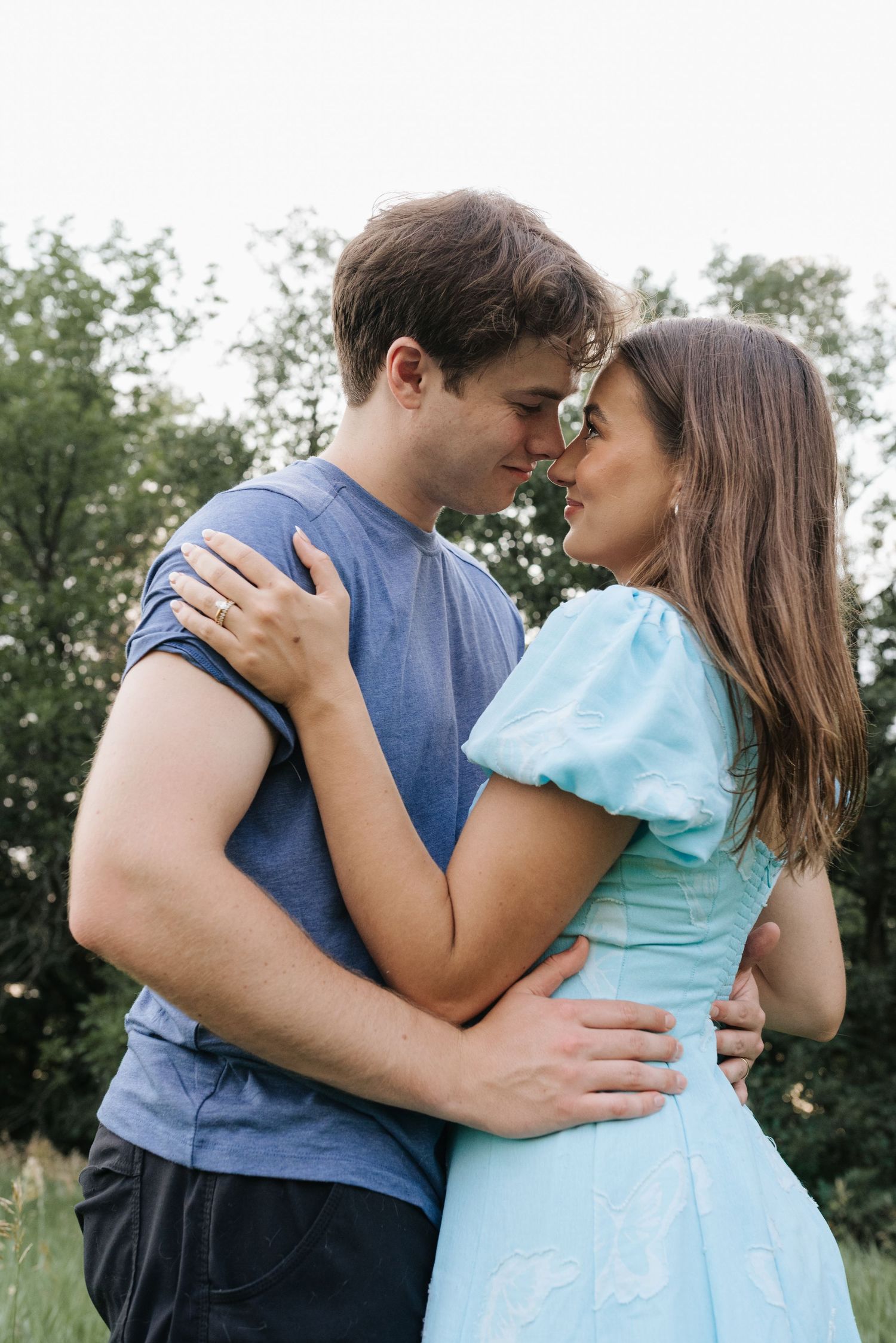 A couple embraces outdoors, nearly kissing, man in blue tee and woman in light blue puff-sleeve dress.