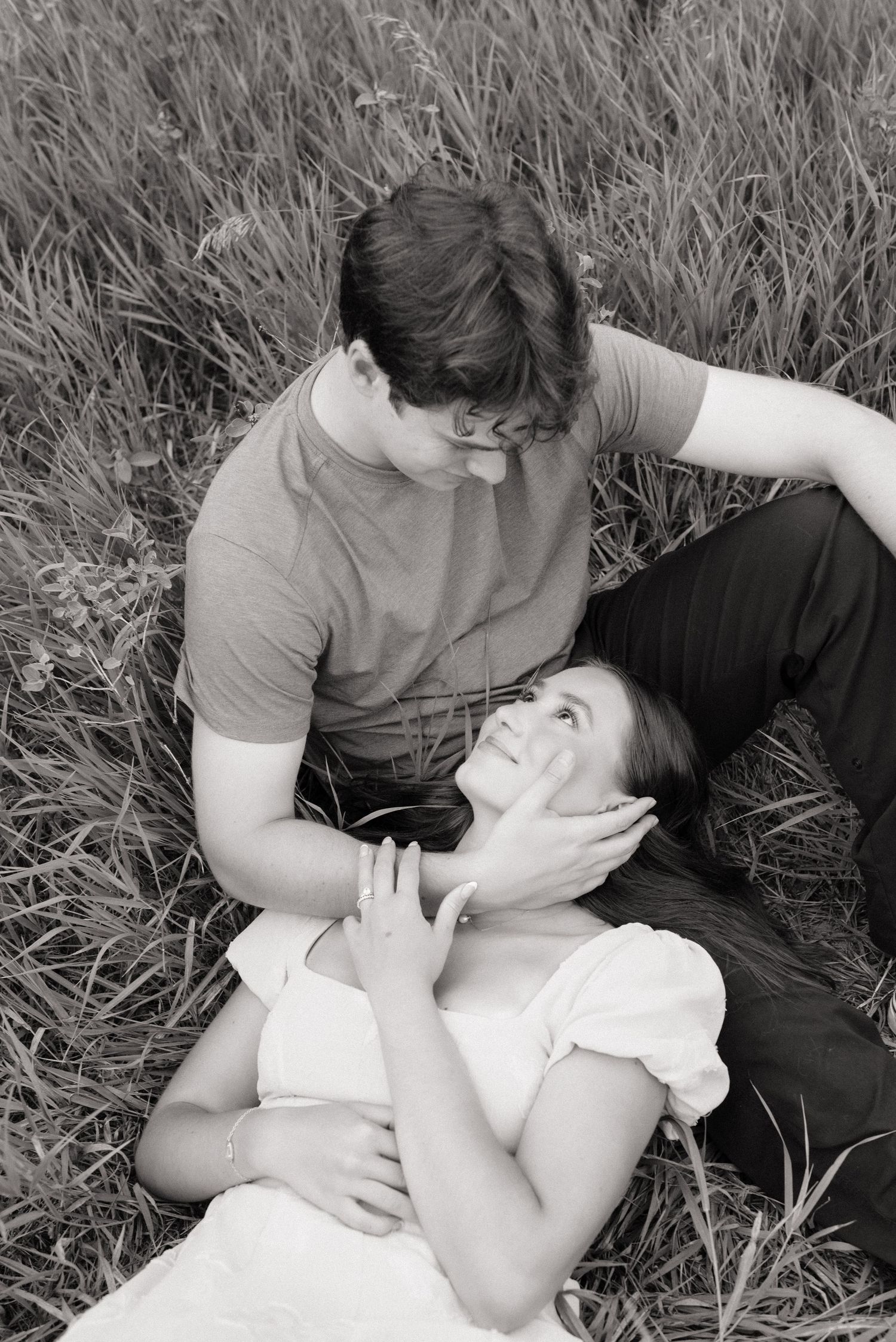 Black and white photo of a romantic couple sitting in a grassy field, woman lying back smiling up at man.