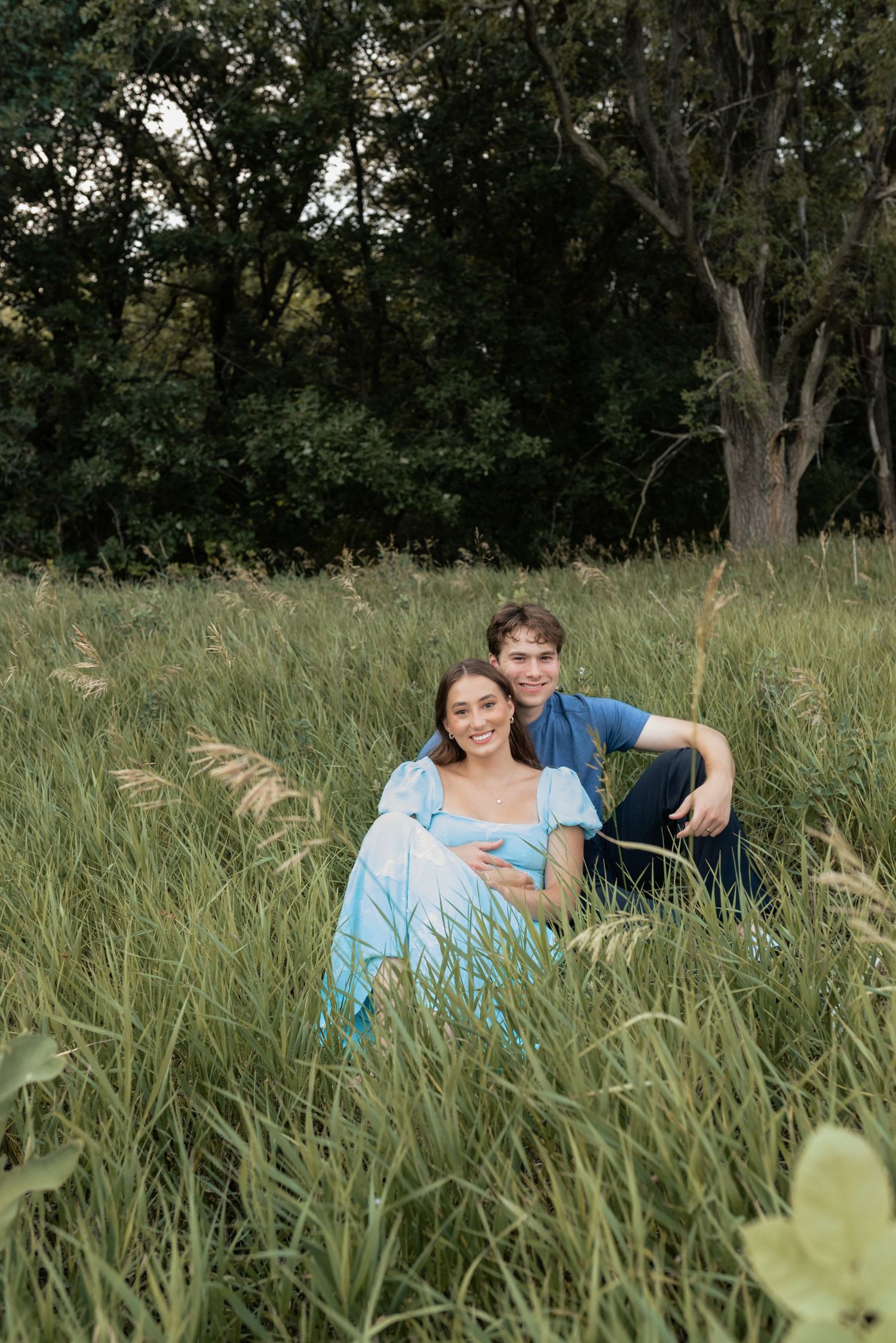 A smiling couple sits together in a lush green meadow, the woman in a light blue dress, surrounded by tall grass and trees.