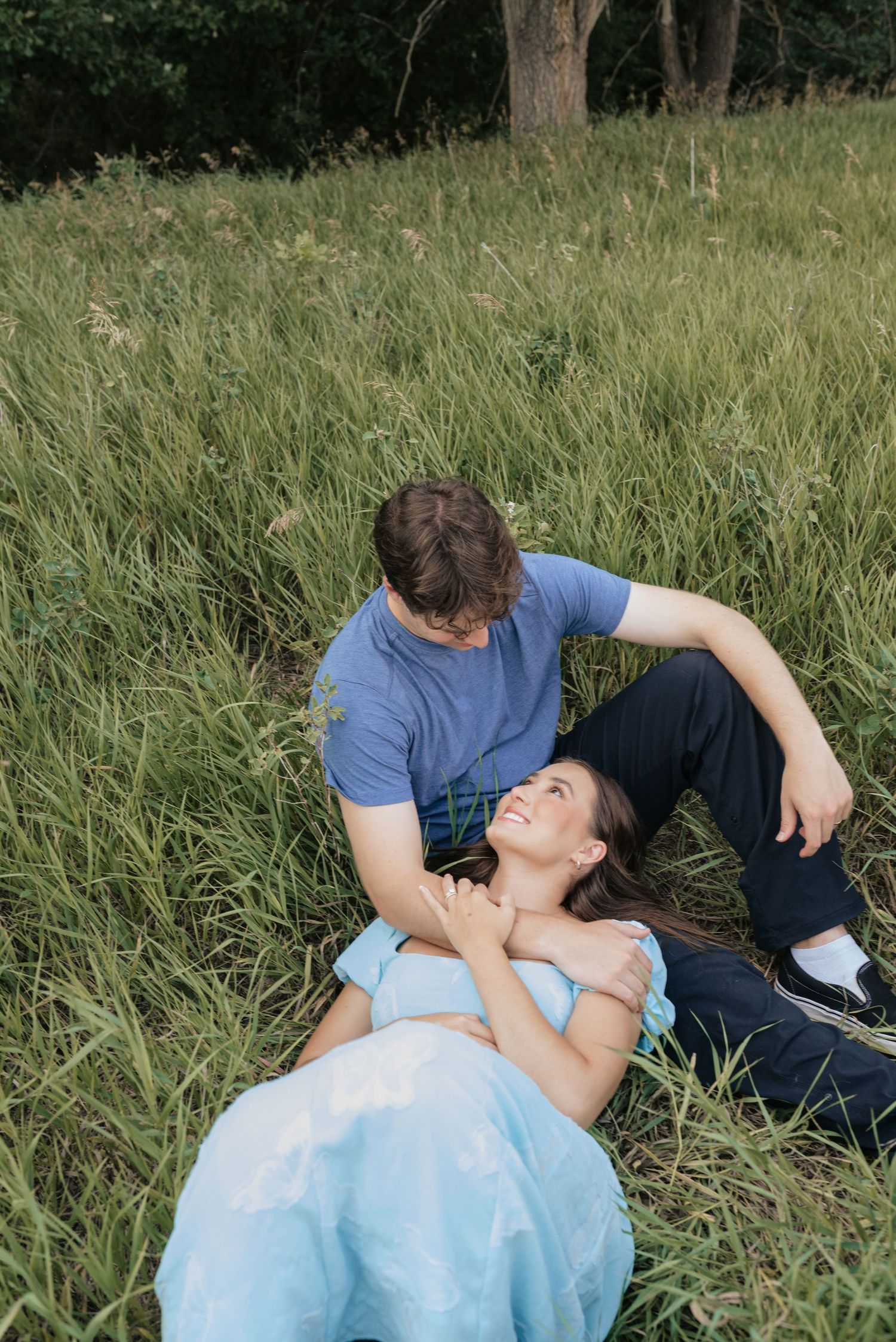 A couple relaxes in a lush green field, woman lying down smiling up at man seated beside her.