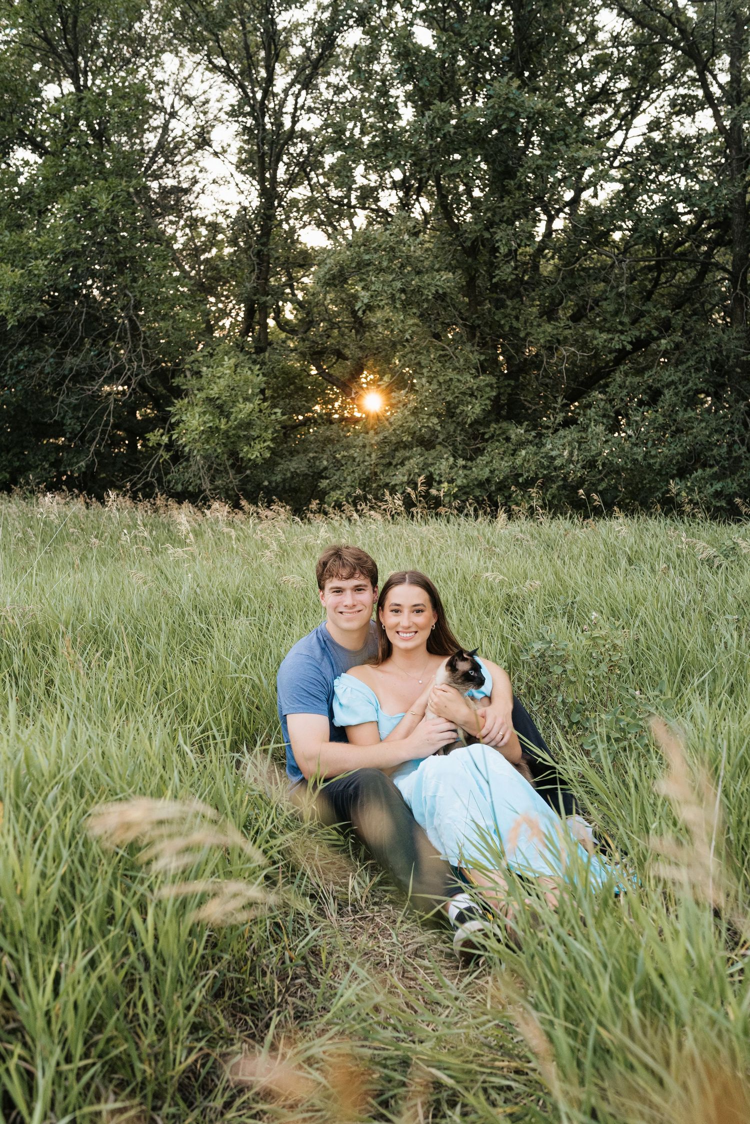 Couple sitting in tall grass field at sunset, smiling and holding a cat, surrounded by lush green trees.