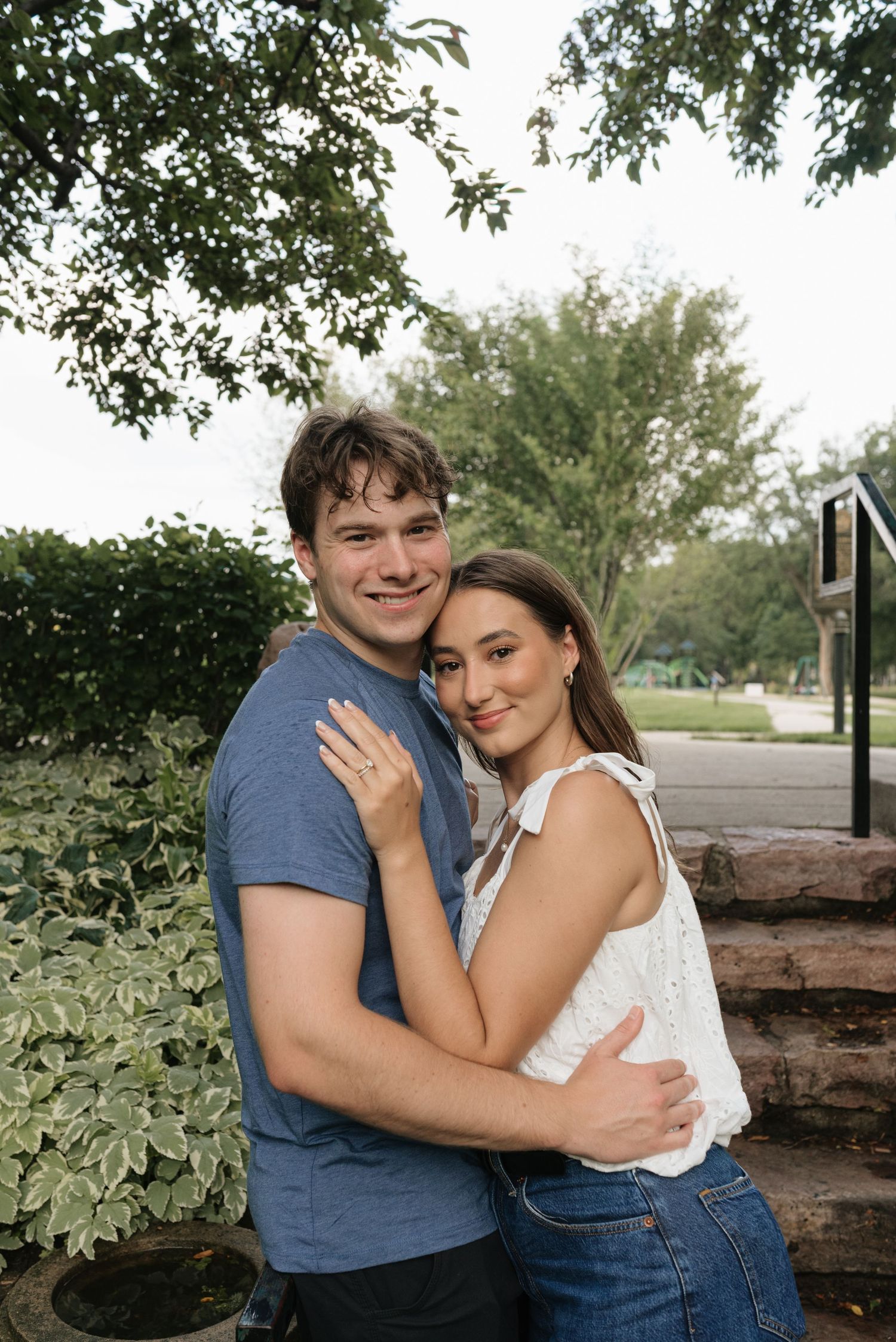 A smiling couple embraces outdoors near lush green shrubs and trees, the woman wearing a white top and jeans.