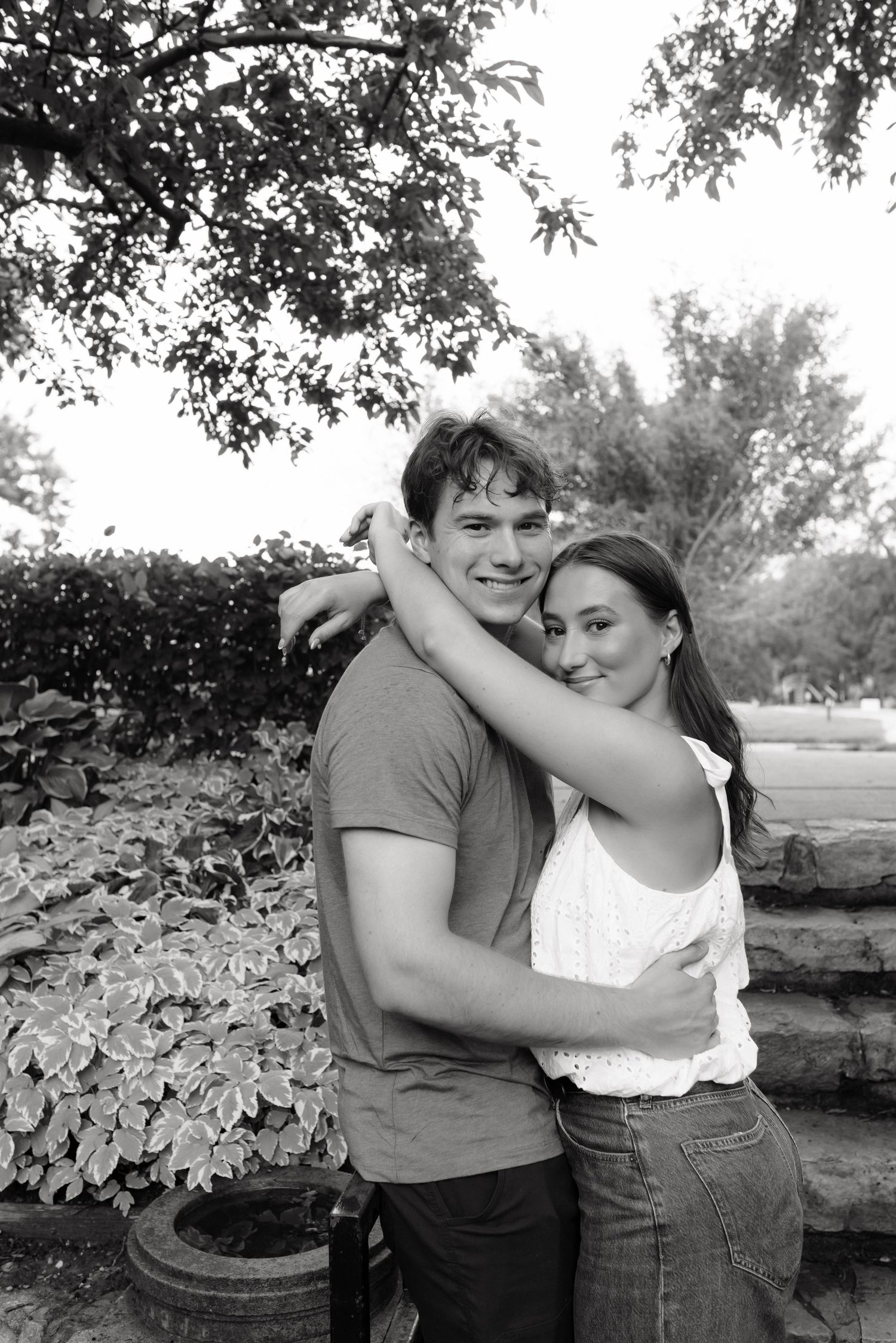 Black and white photo of a young couple embracing outdoors, woman wrapping arms around mans neck, smiling together.