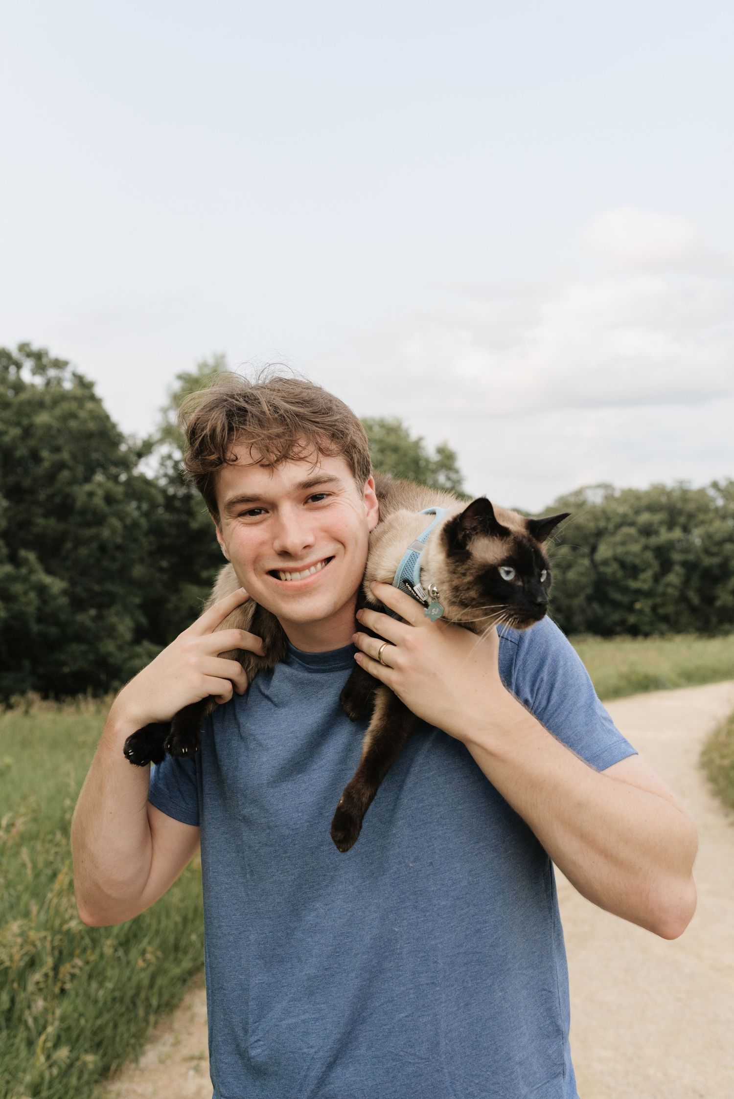 Smiling young man in blue t-shirt holding a Siamese cat on his shoulders outdoors on a dirt path.