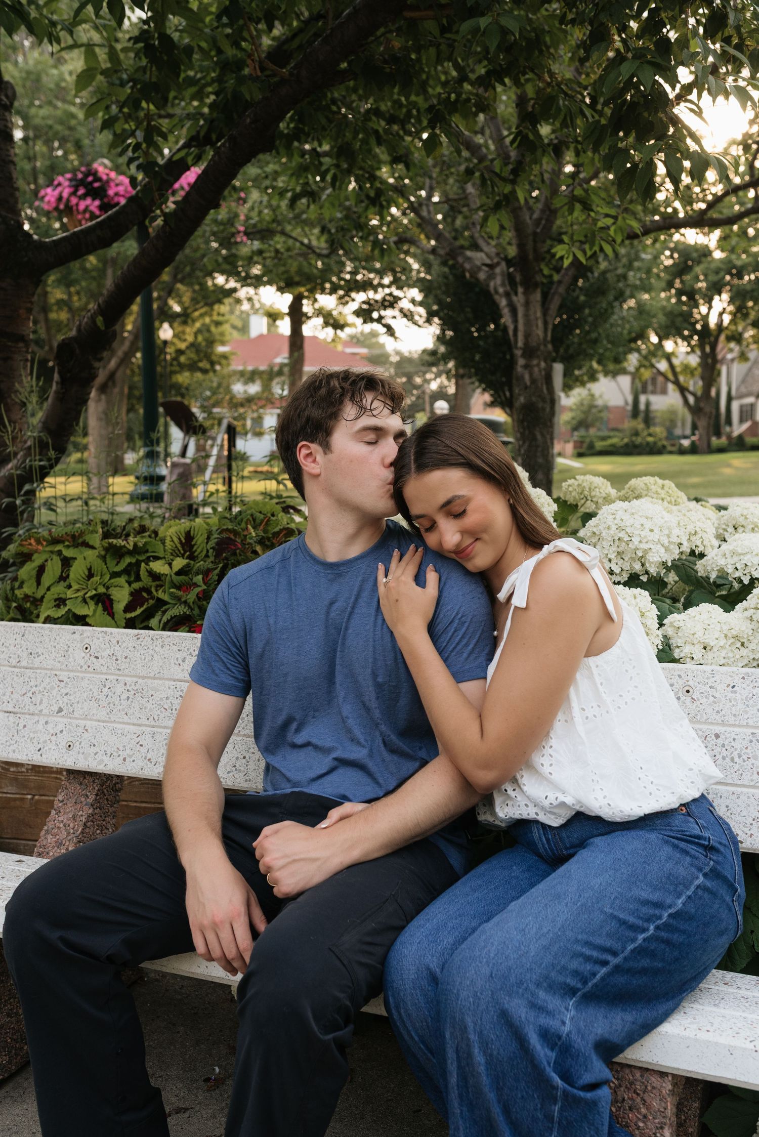 A couple sits on a white bench in a park, the man kissing the woman's head as she leans on his shoulder.