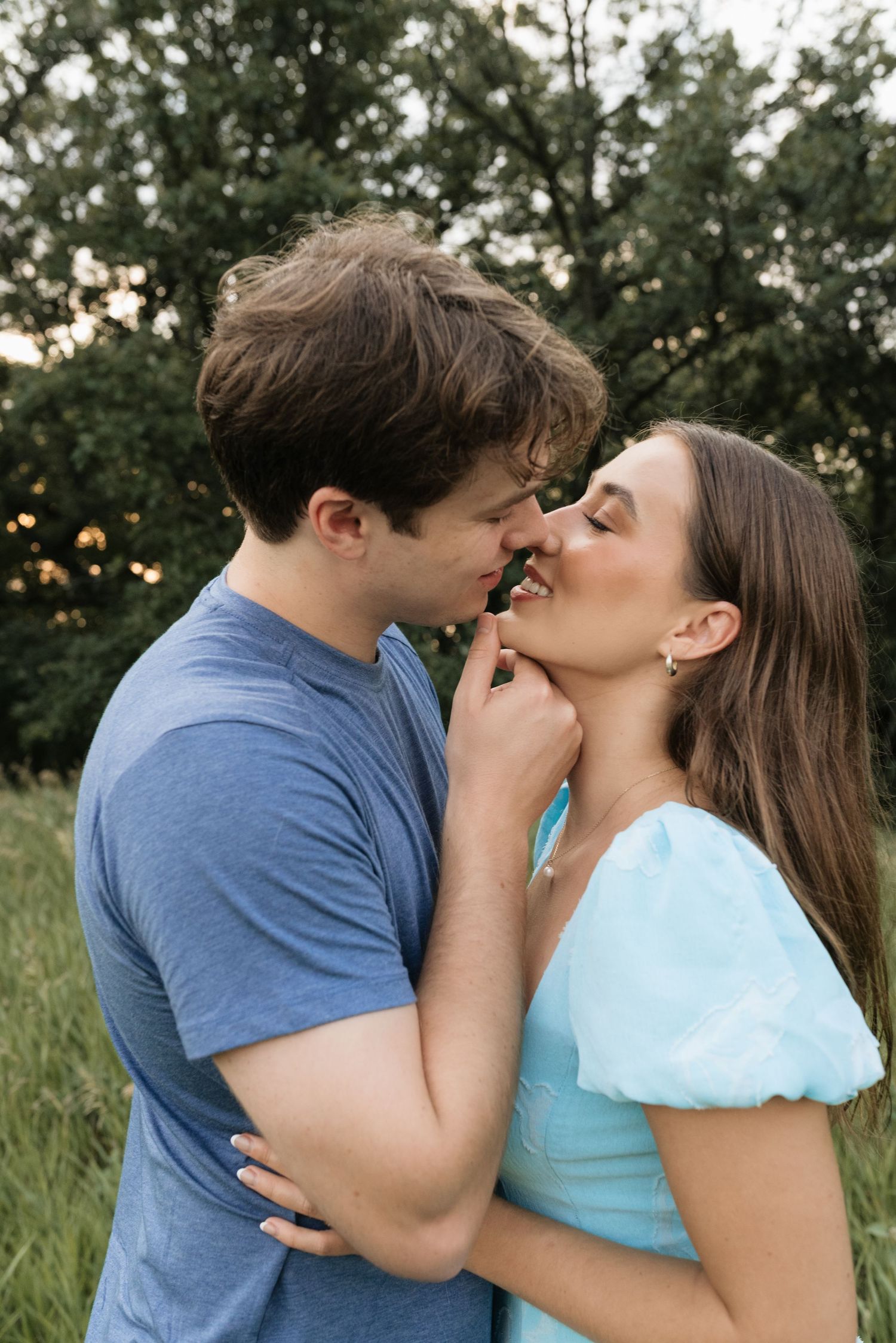 A couple shares a romantic moment outdoors, the man in blue gently holding the woman's chin as they lean in to kiss.