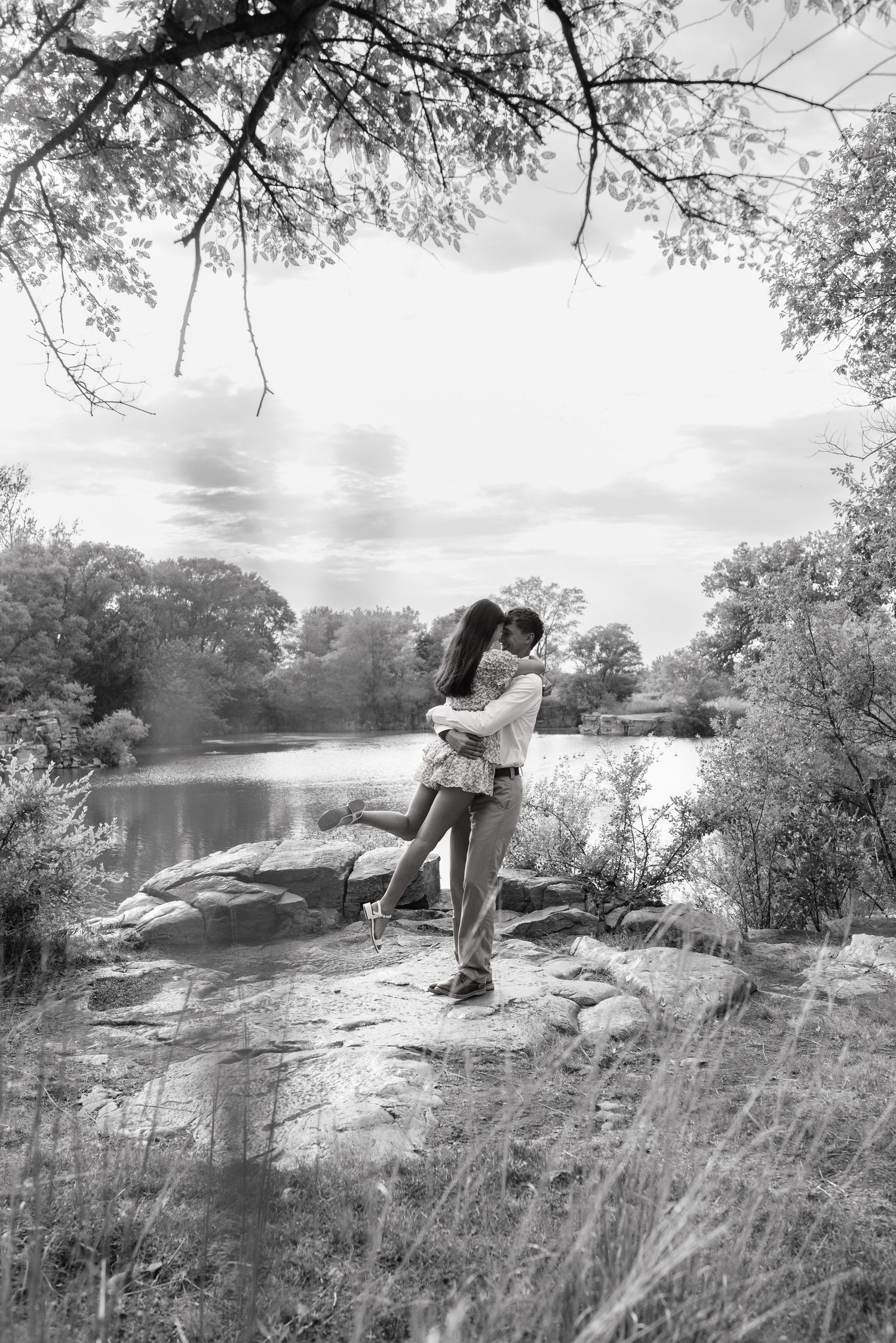 Black and white photo of a couple embracing and kissing on rocks by a serene lake surrounded by lush trees.