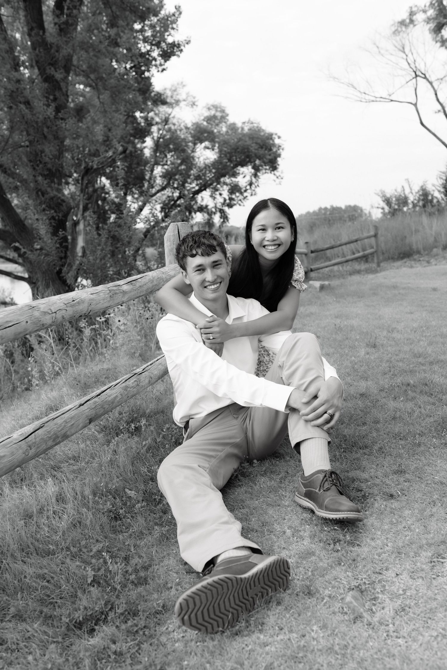 Black and white photo of happy couple sitting against fence on grass, woman hugging man from behind.