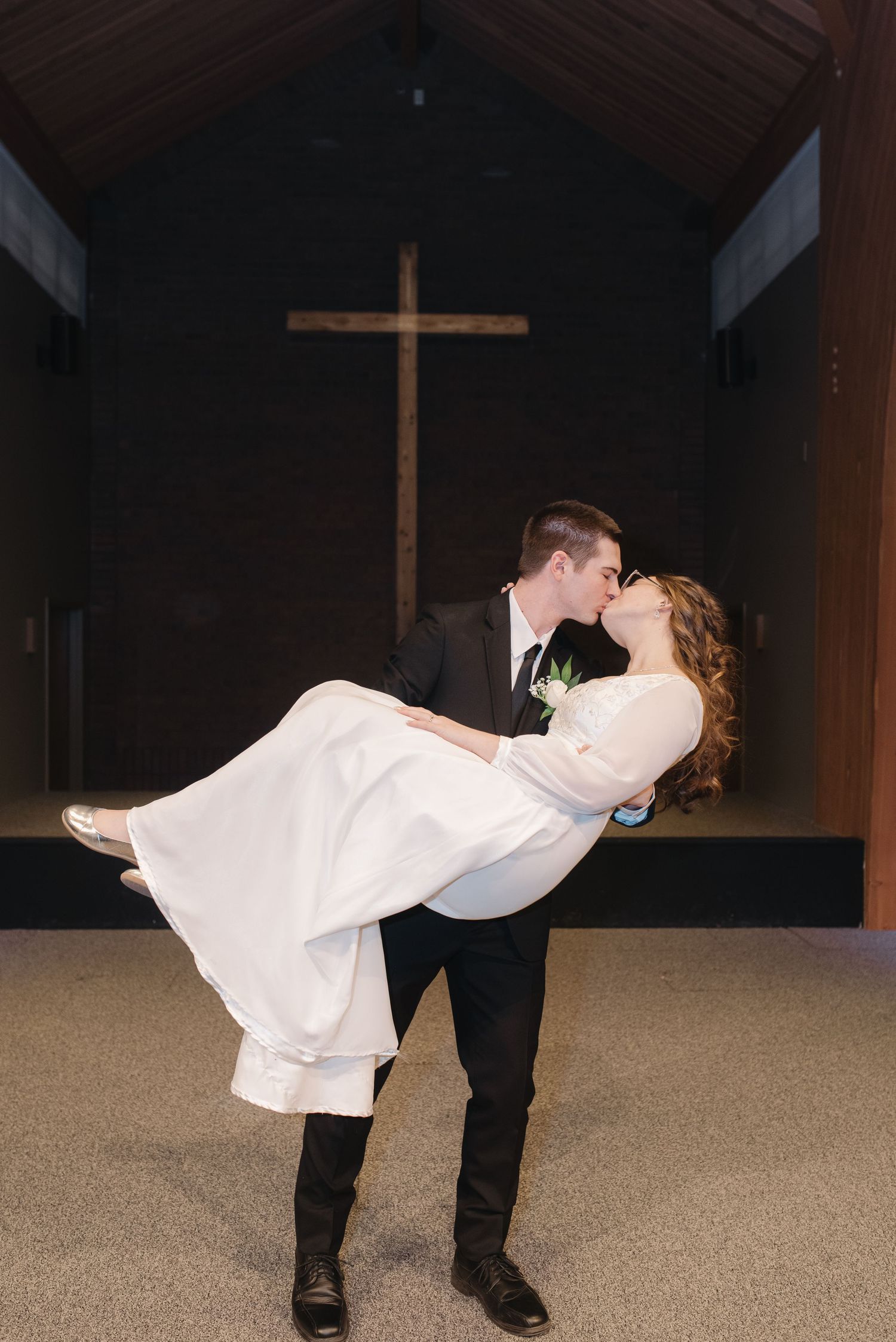 A groom lifts his bride in a white dress for a kiss in front of a wooden cross in a church.