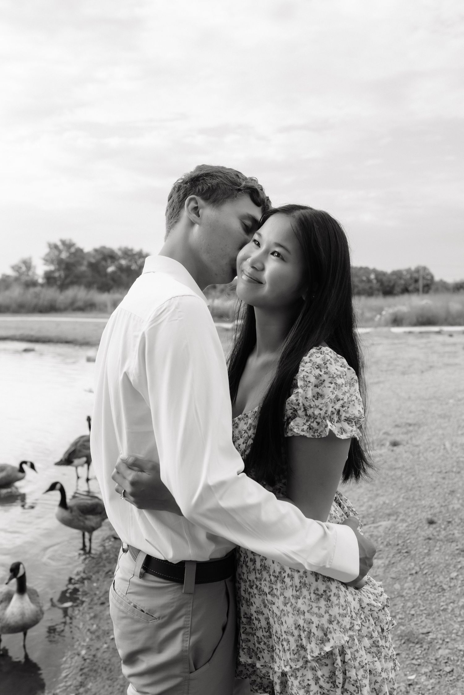 Black and white photo of a couple embracing near a lake with geese in the background.