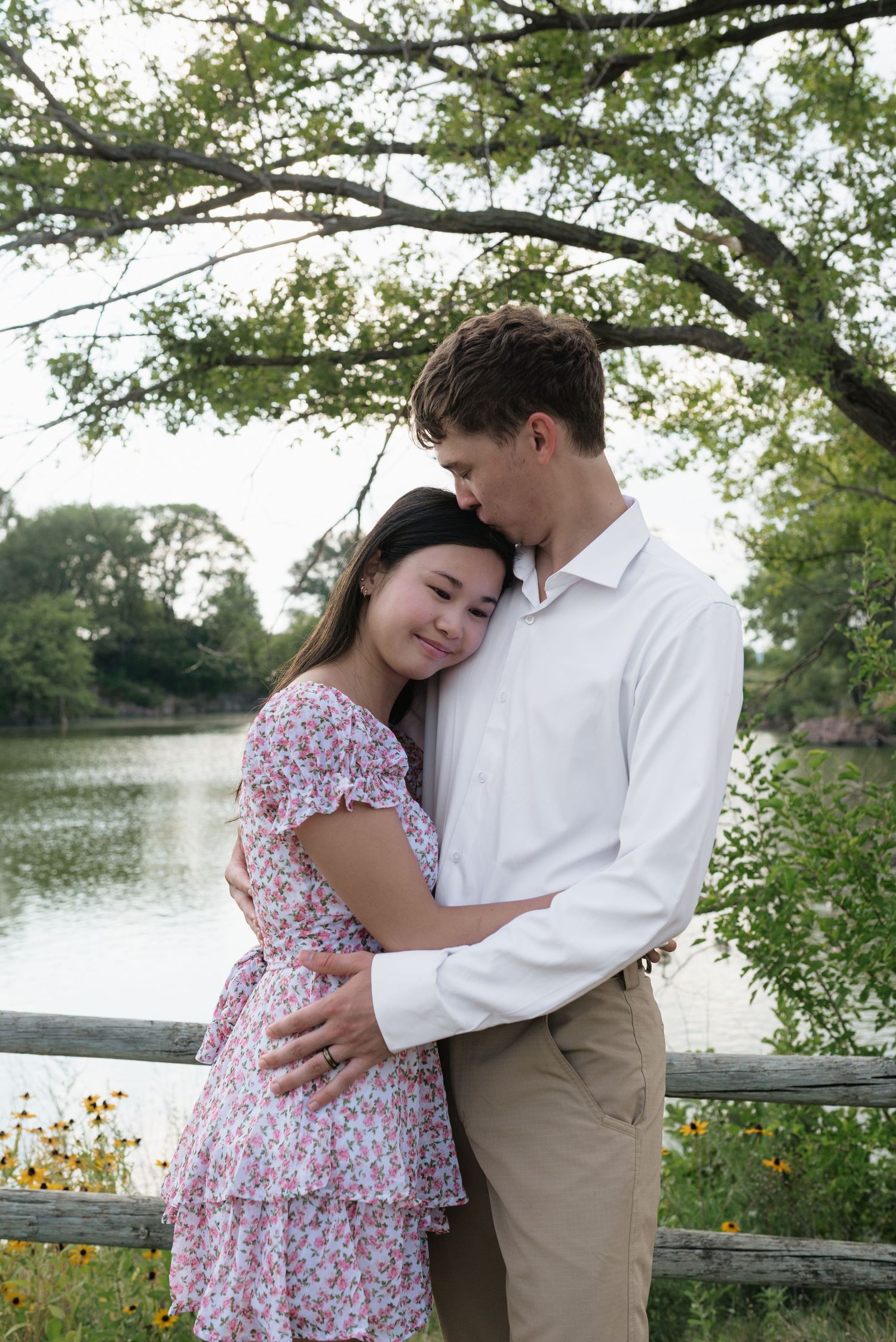 Couple embracing tenderly by a wooden fence near a serene pond with lush trees.