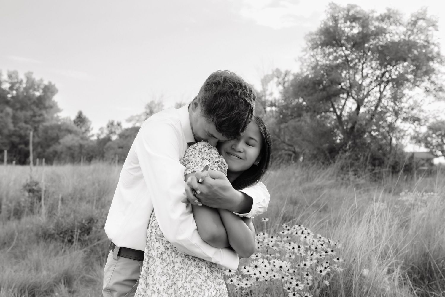 Black and white photo of a couple sharing a tender embrace in an open field with tall grass.