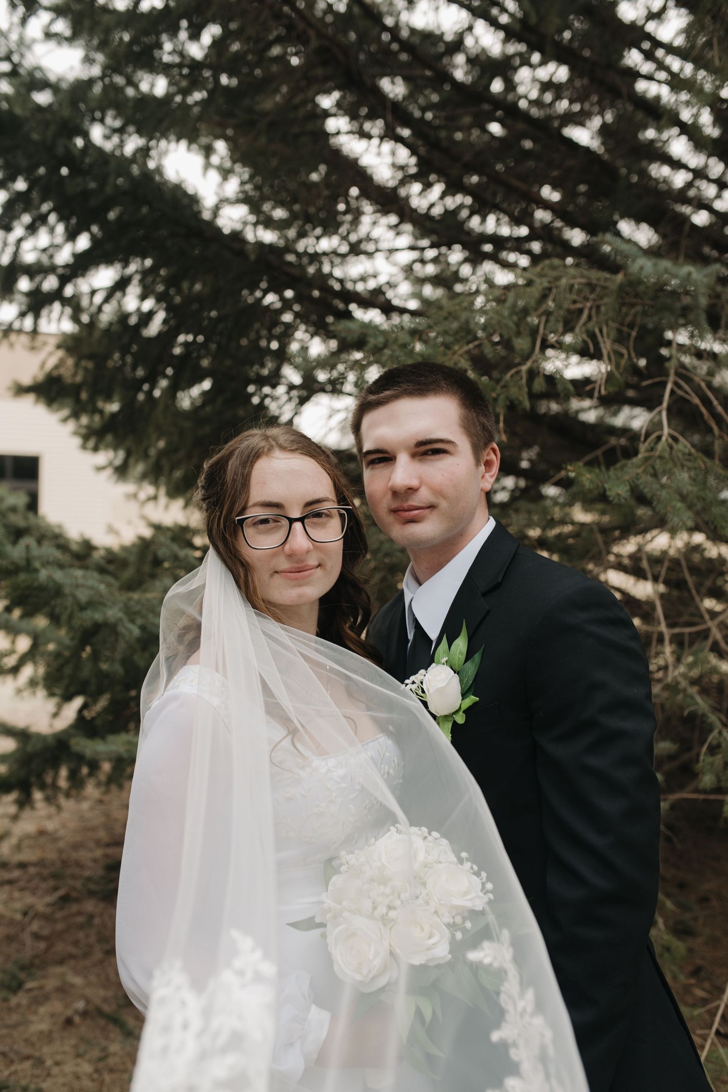 Bride in white gown with veil and groom in black suit pose outdoors near evergreen tree.
