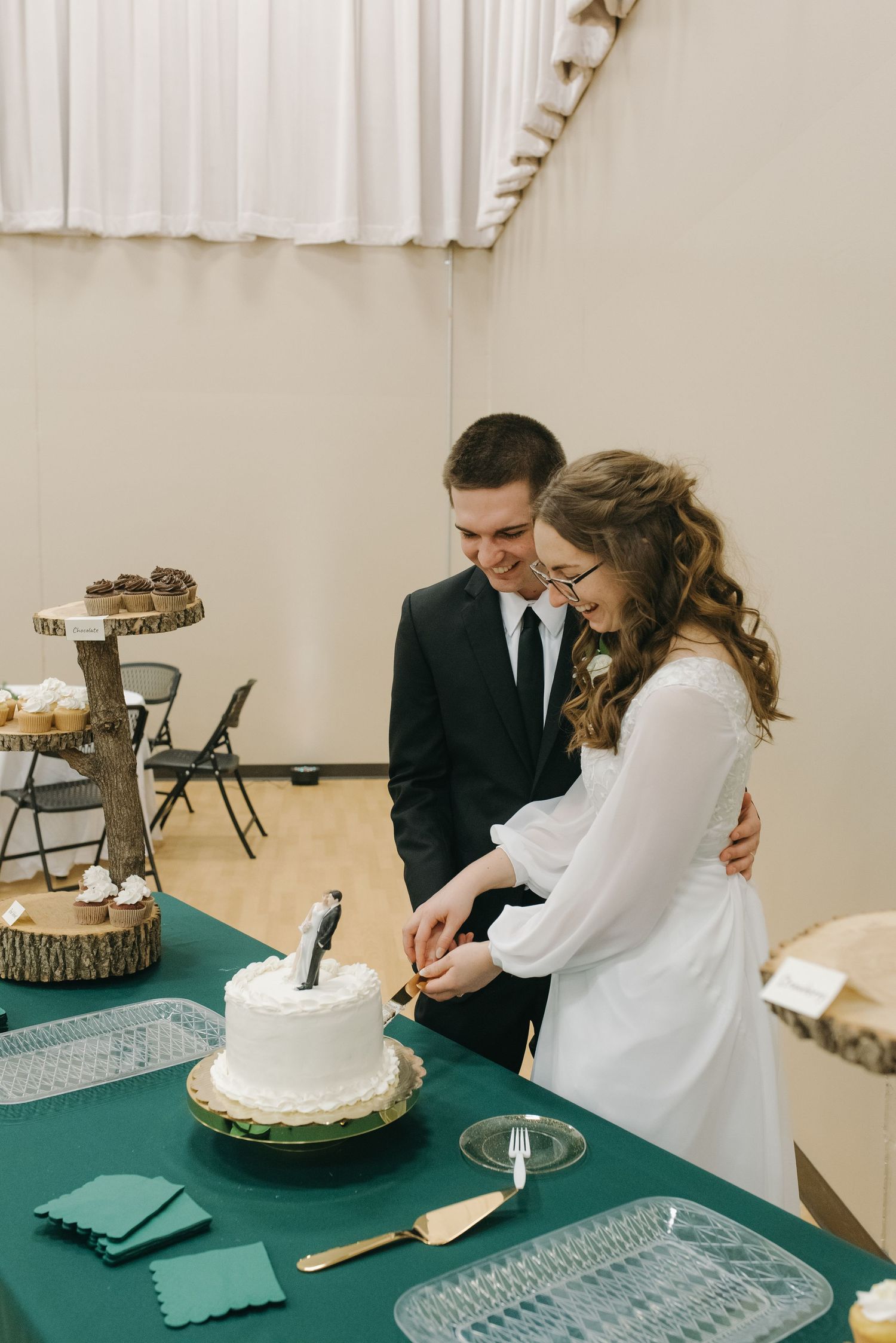 Smiling newlyweds cutting white wedding cake together at green-draped reception table with desserts.