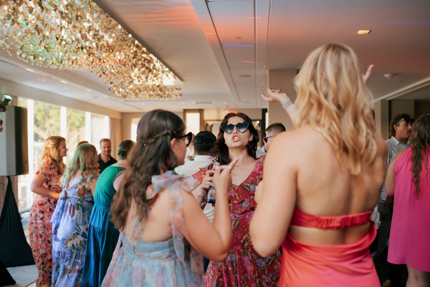 Guests in colorful dresses dance and celebrate beneath elegant chandelier at wedding reception.