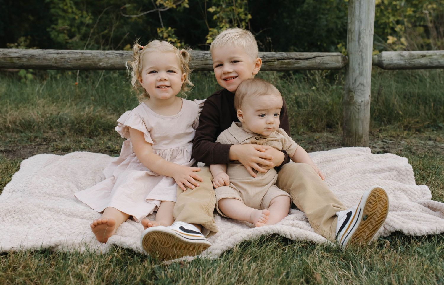 Three blonde siblings sitting together on a white blanket in a grassy outdoor setting near a wooden fence.