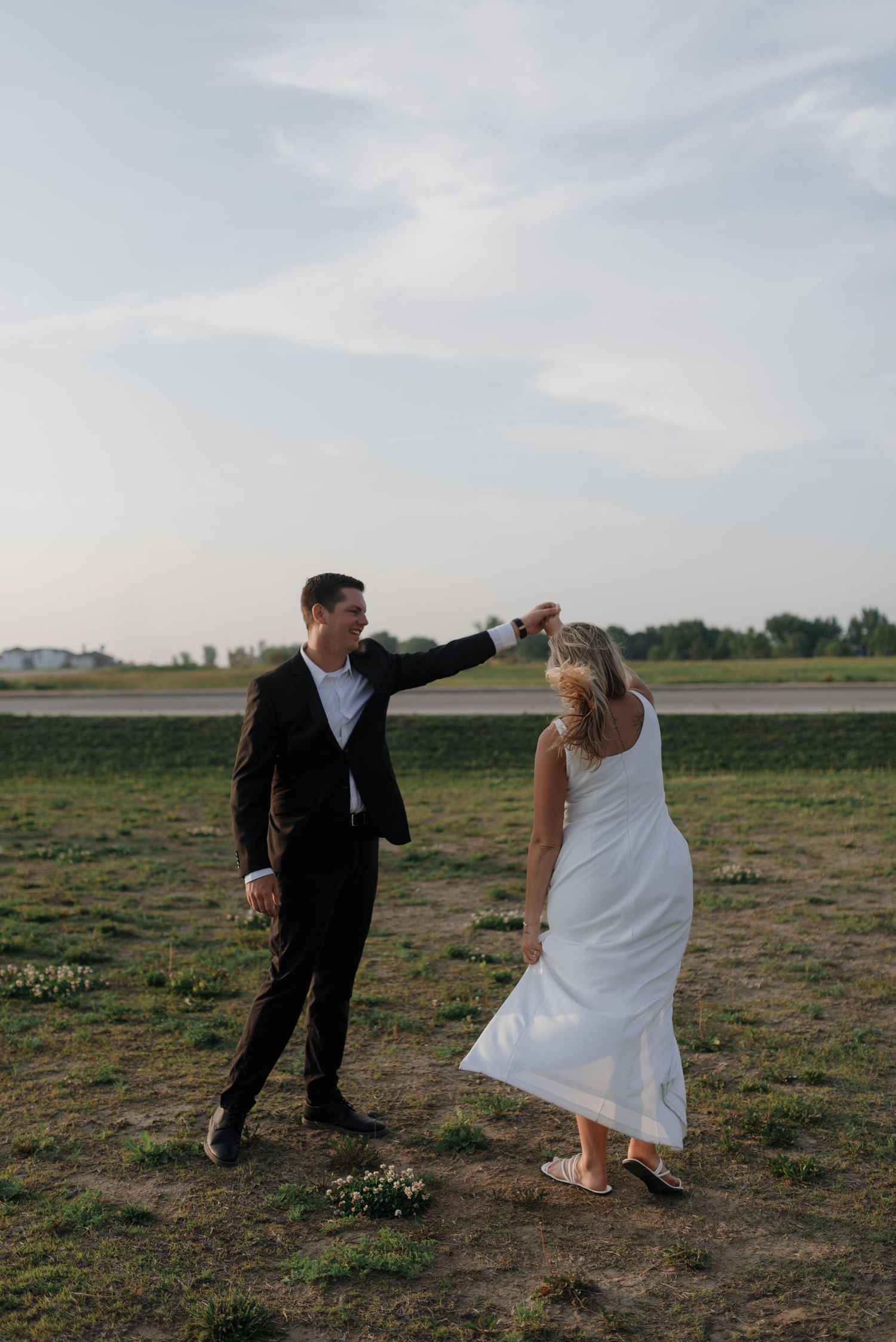 Groom twirling bride in white dress outdoors in open field during golden hour engagement shoot.