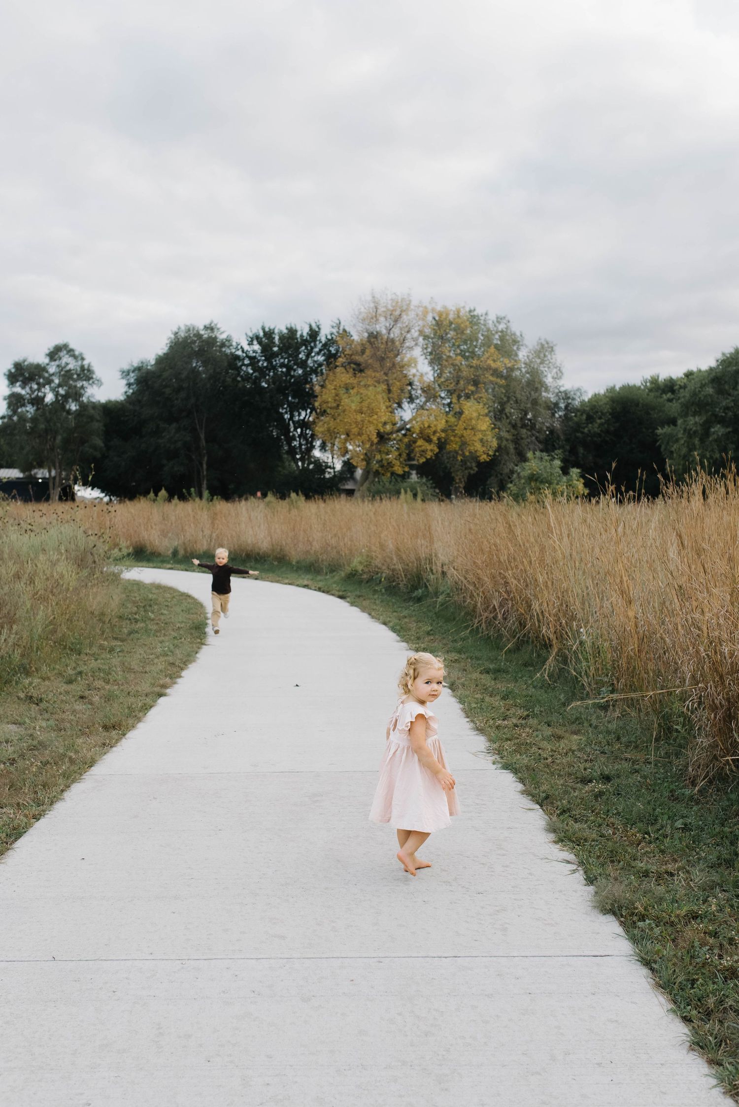 Two young children walking barefoot on a winding concrete path surrounded by tall golden grass.