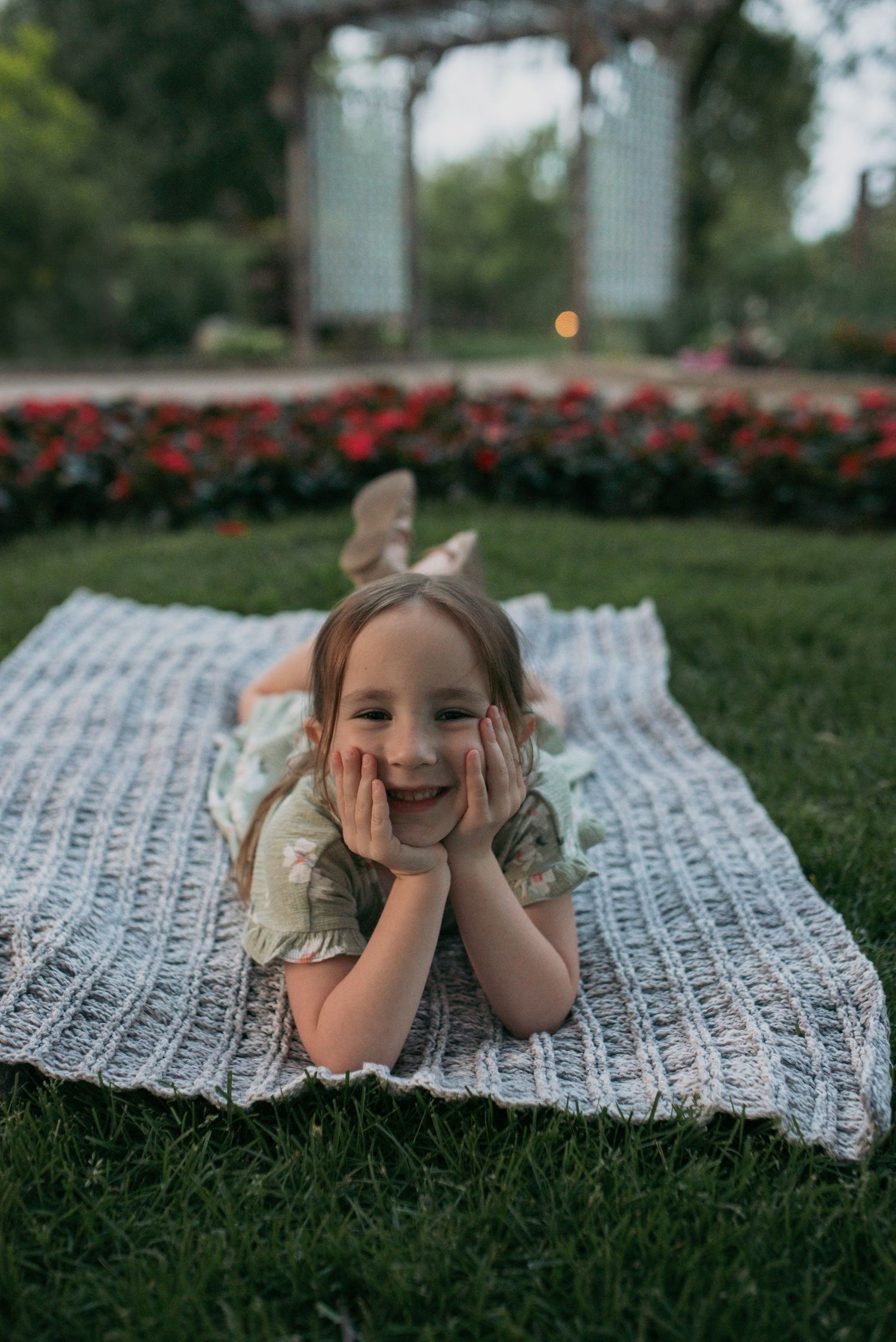 A cheerful young girl resting on a knit blanket in a garden, smiling with hands on cheeks.