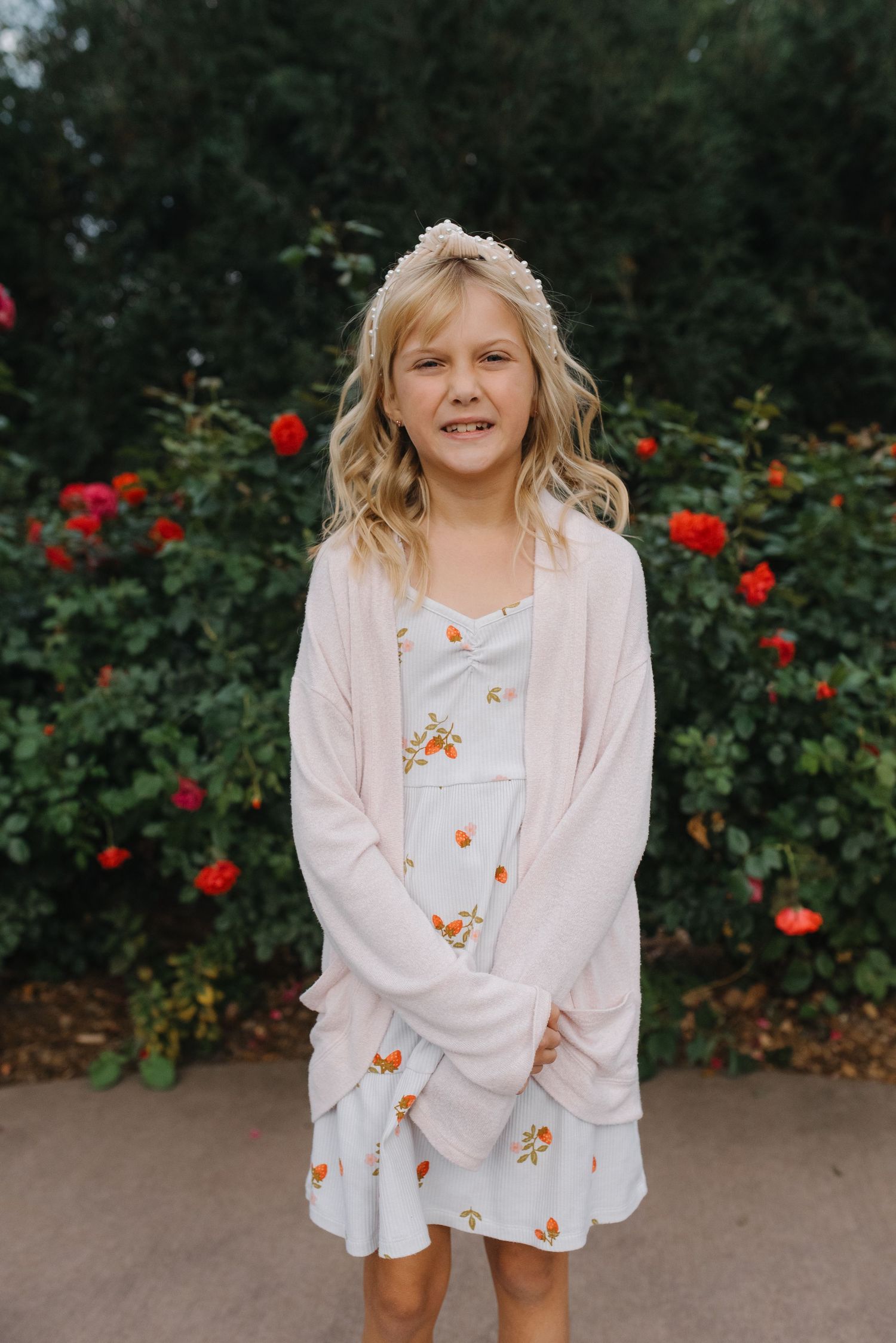 Smiling blonde girl wearing a floral dress and pink cardigan posing in front of blooming red roses.