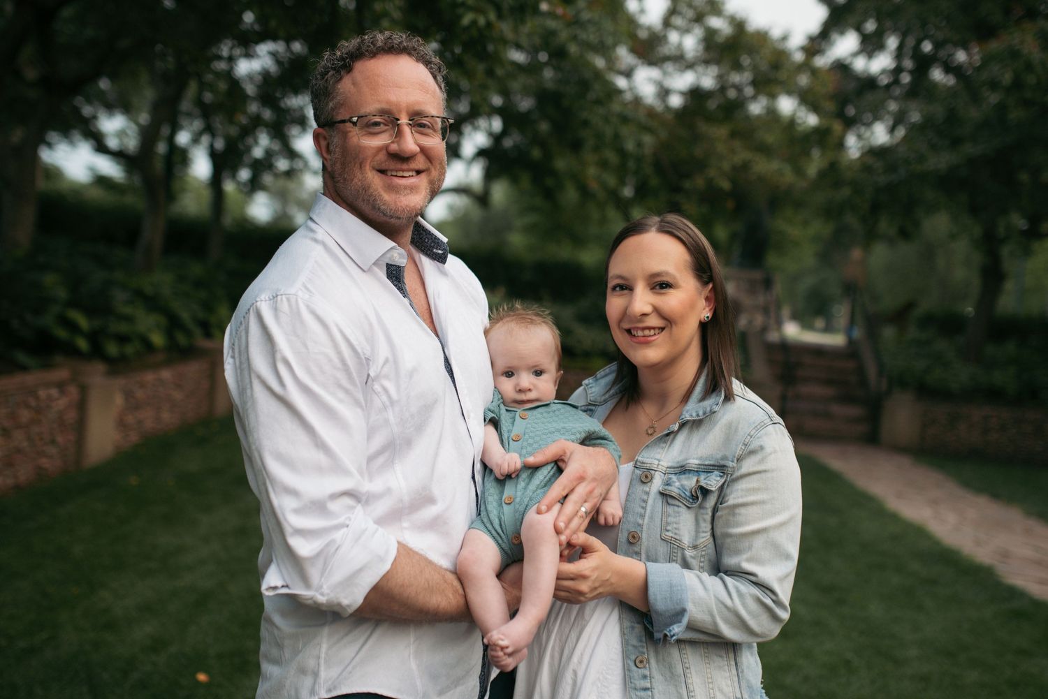 Smiling couple holds baby outdoors in green park with trees in background.