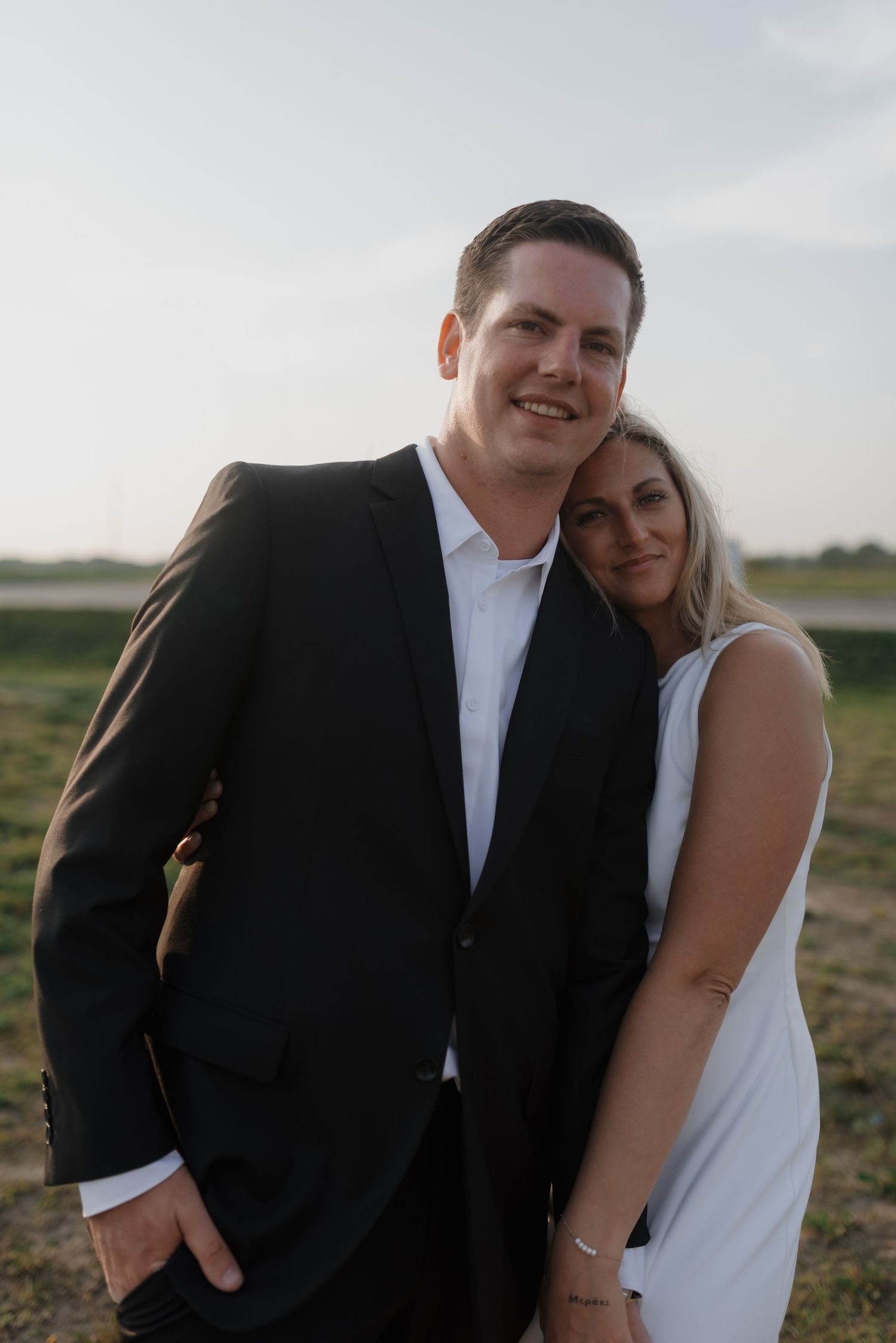 Couple posing together in field at dusk, man in black blazer and woman in sleeveless white dress.